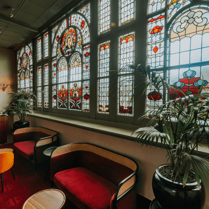 Ornate stained glass windows illuminate a cozy lounge with red sofas and green plants.
