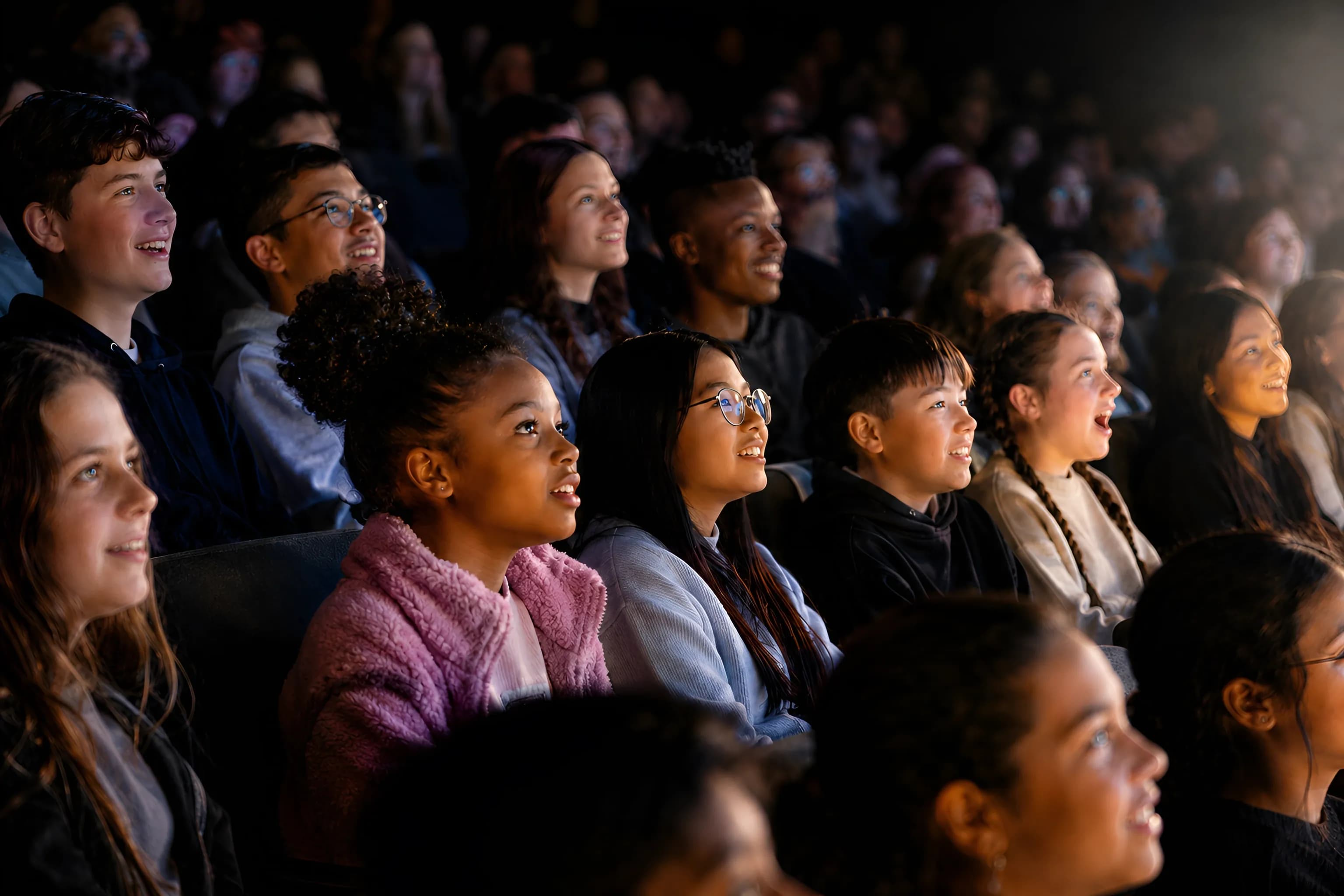 Diverse young students watch intently from an audience, faces illuminated by light.
