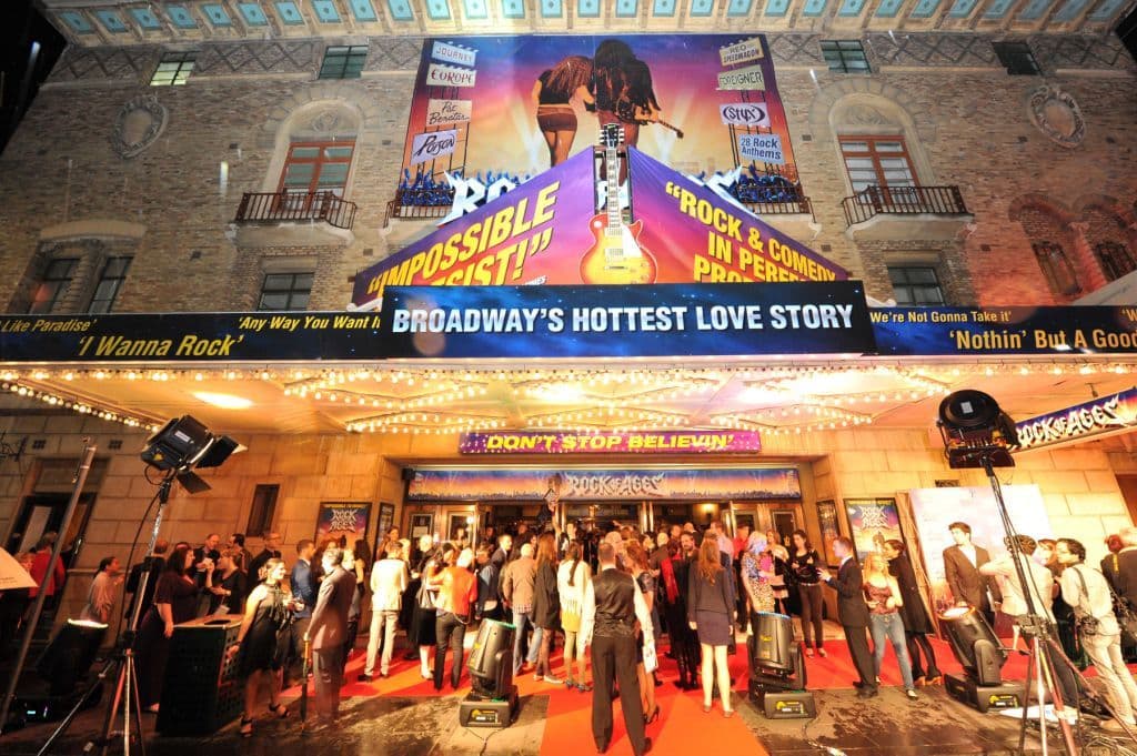 Crowd gathers outside Broadway's Rock of Ages theater under bright lights at night.