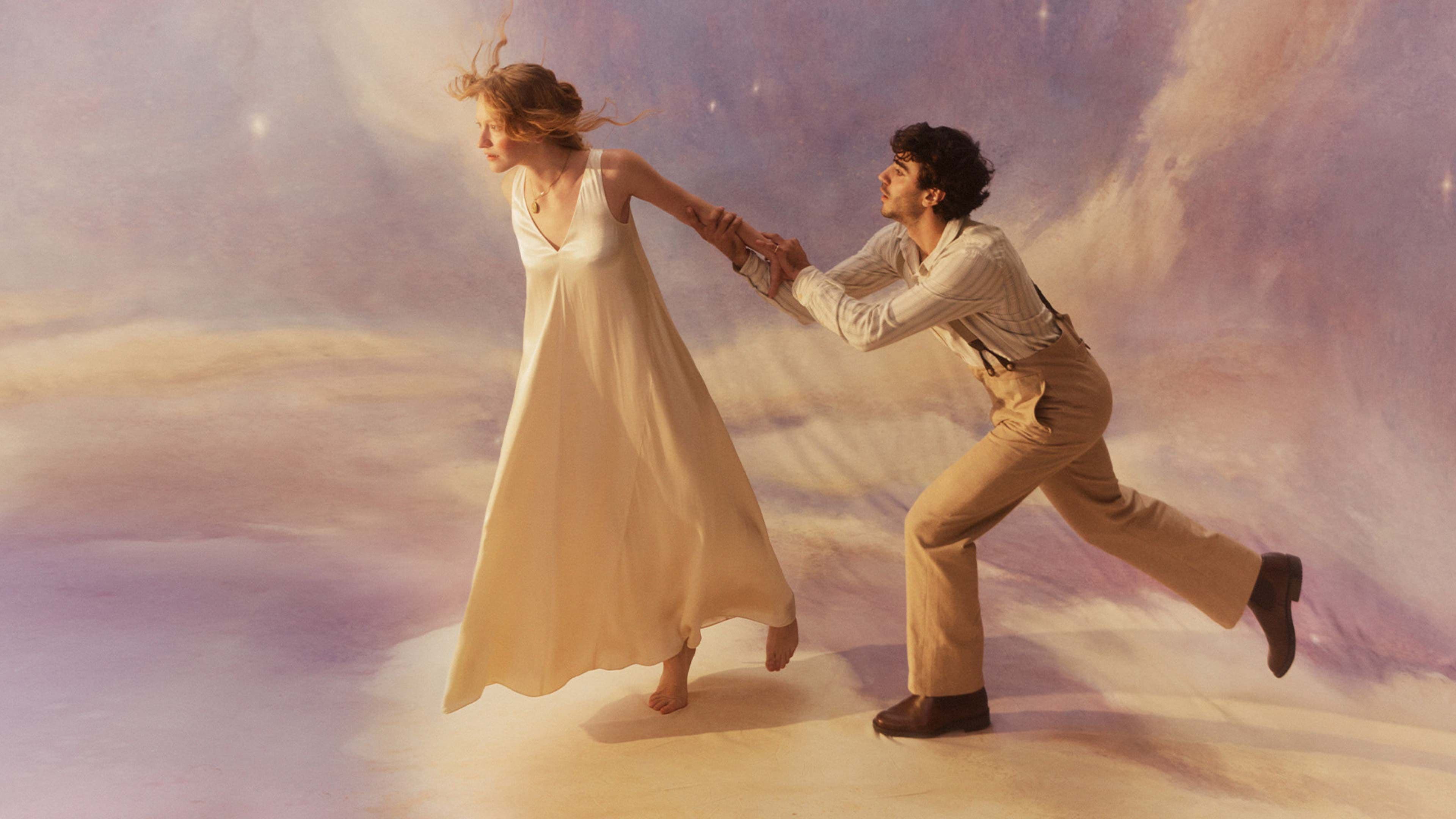 Woman in white dress leading man in tan clothes across dreamy, starry clouds.
