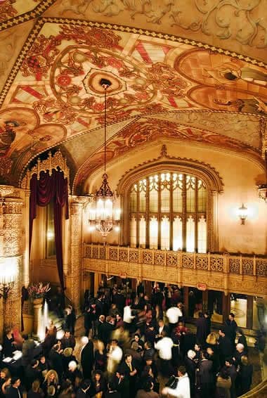 Ornate ballroom with a detailed painted ceiling, arched windows, and a crowd.
