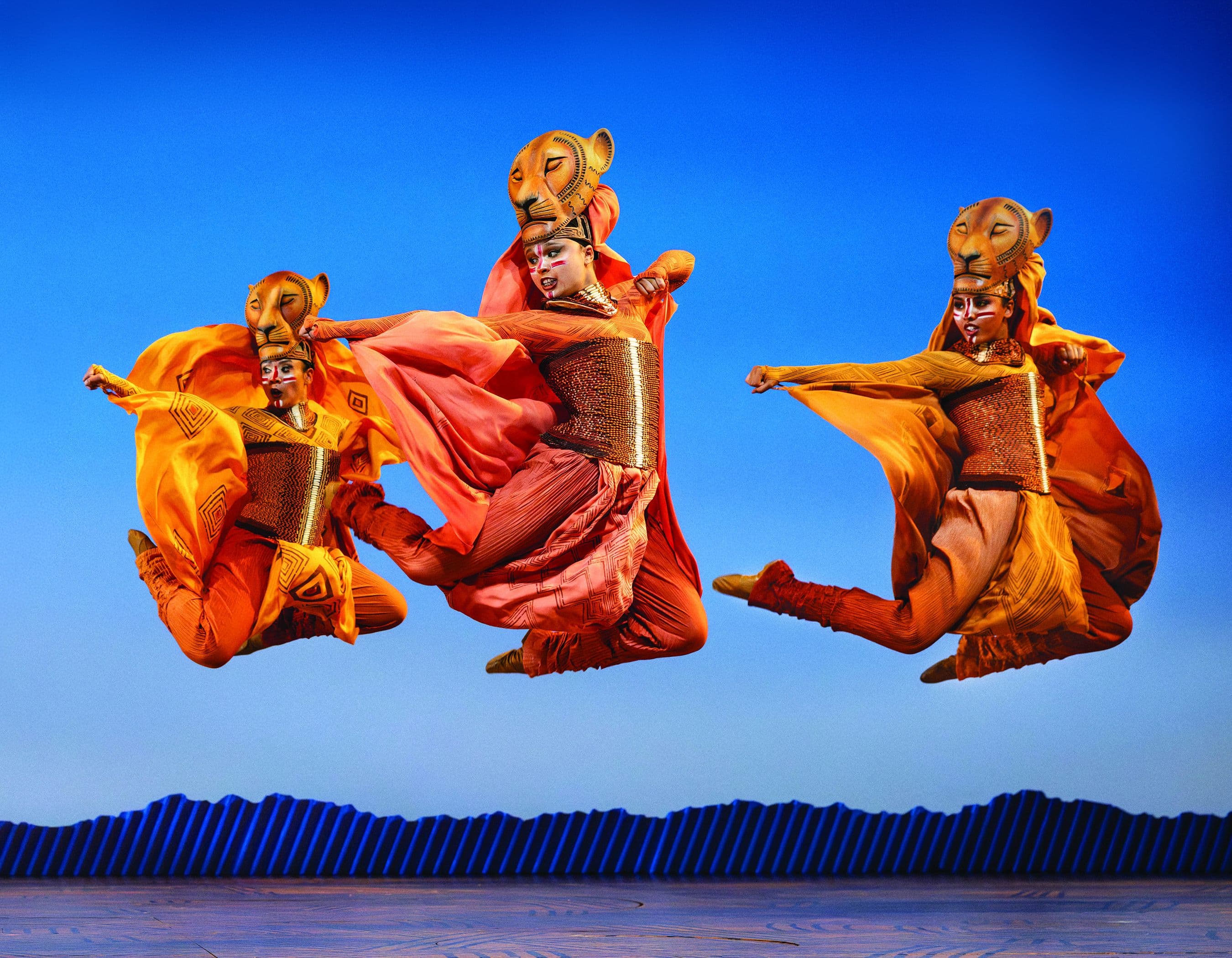 Three performers in vibrant, animal-inspired costumes jump against a blue sky.