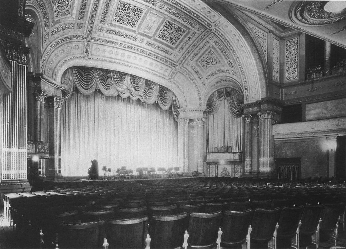 Black and white image of an empty theatre stage with ornate curtains and seating area.