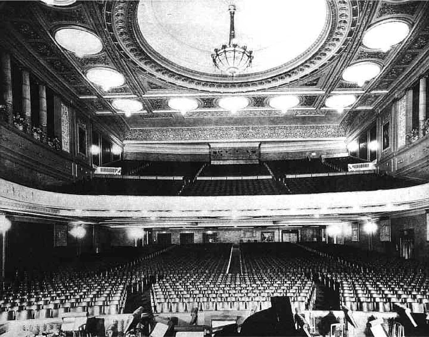 Ornate, empty theatre interior with rows of seats, balcony, and a large chandelier.