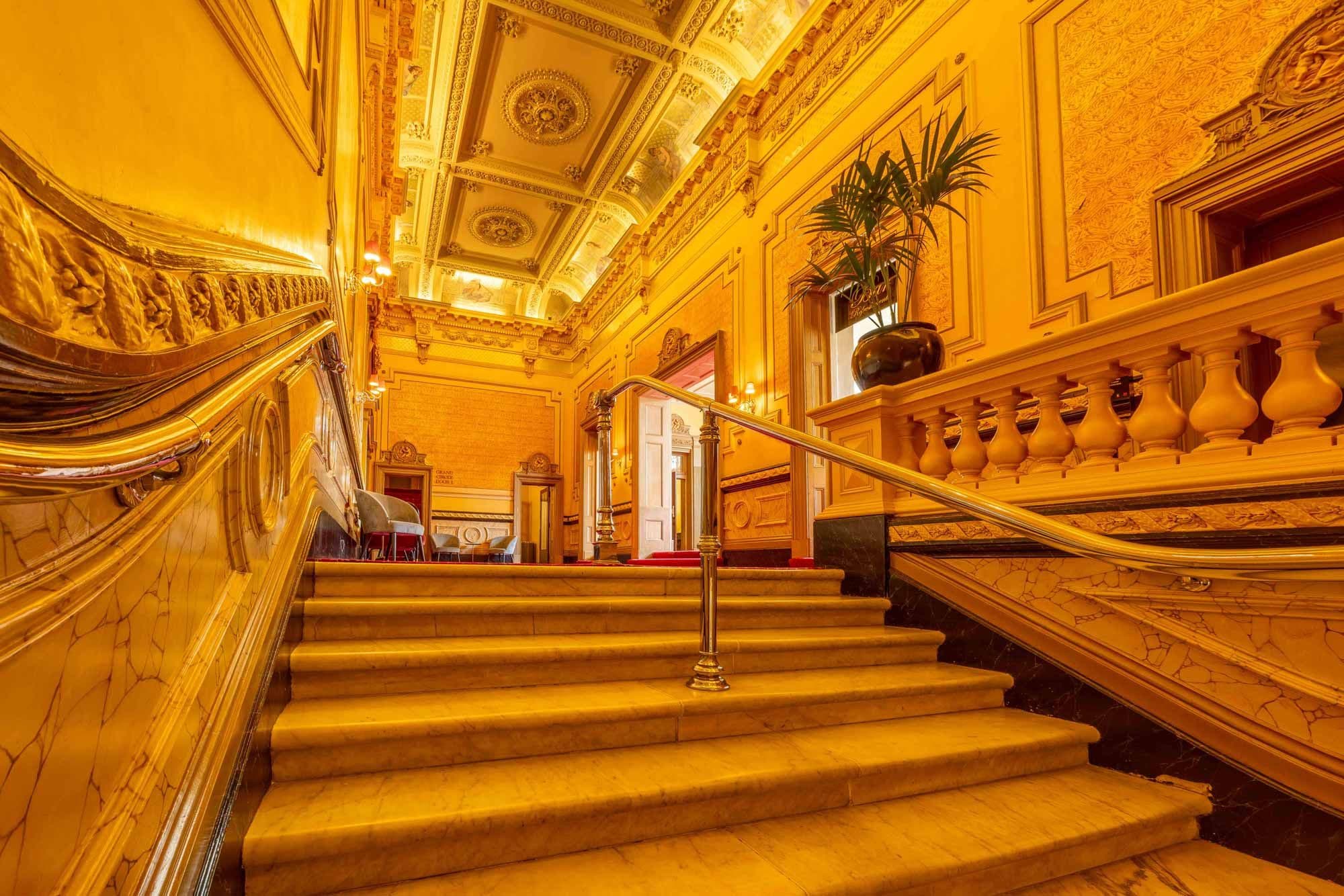 Ornate golden staircase with elaborate railings and decorative ceiling in a luxurious building.