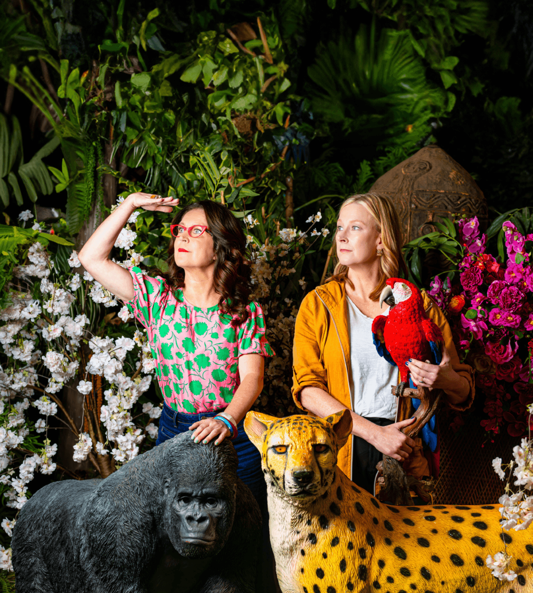 Two women with a parrot and lifelike animal statues in a vibrant jungle setting.