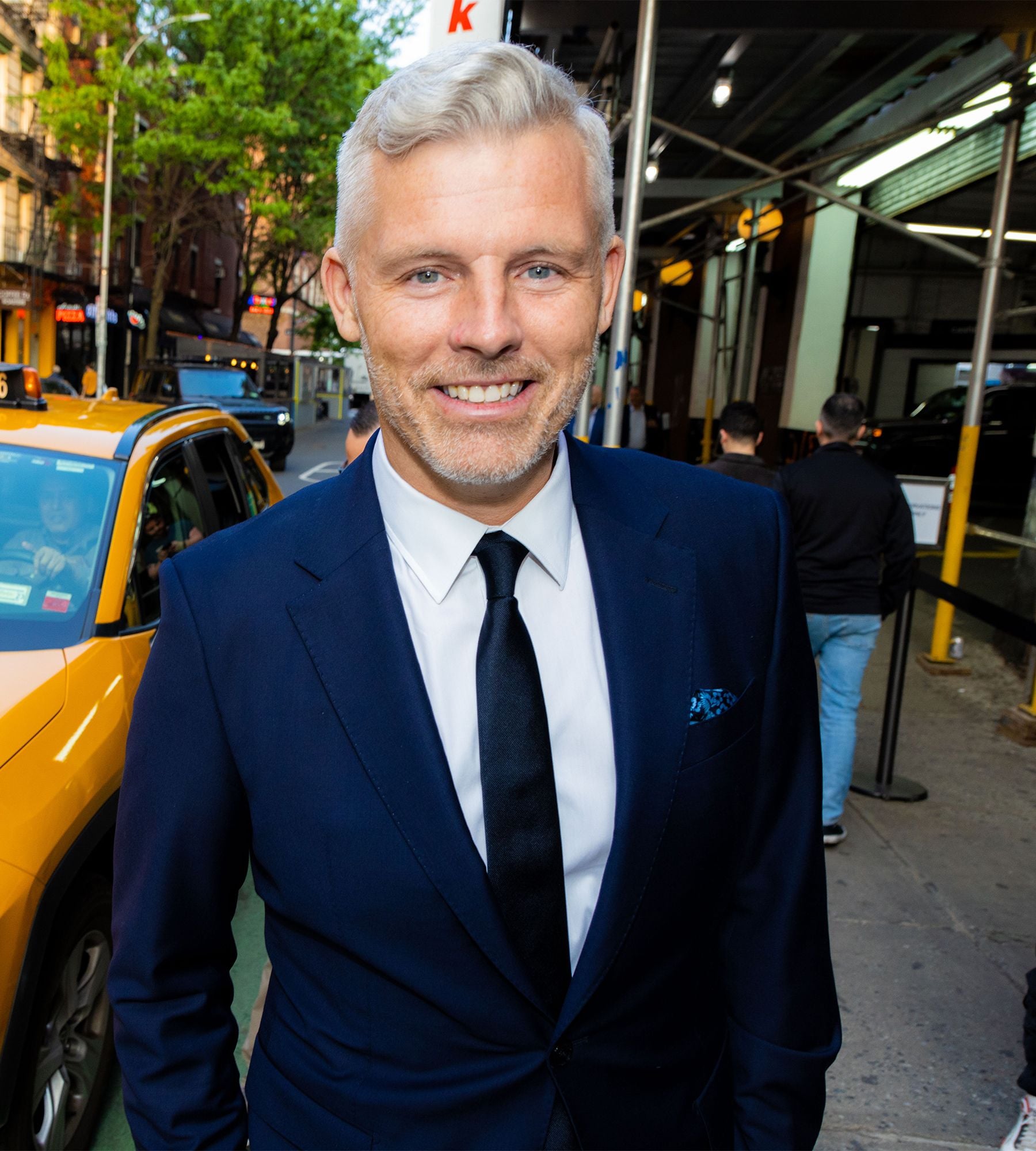 Smiling man in blue suit and black tie standing on city street near a taxi.