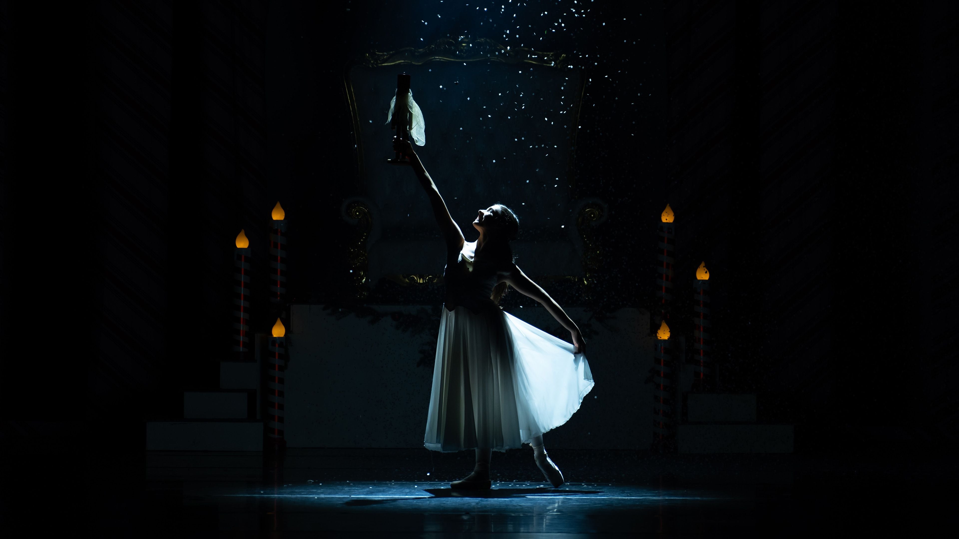 Dancer in a white dress with raised arm and hat, silhouetted against dim candlelight.