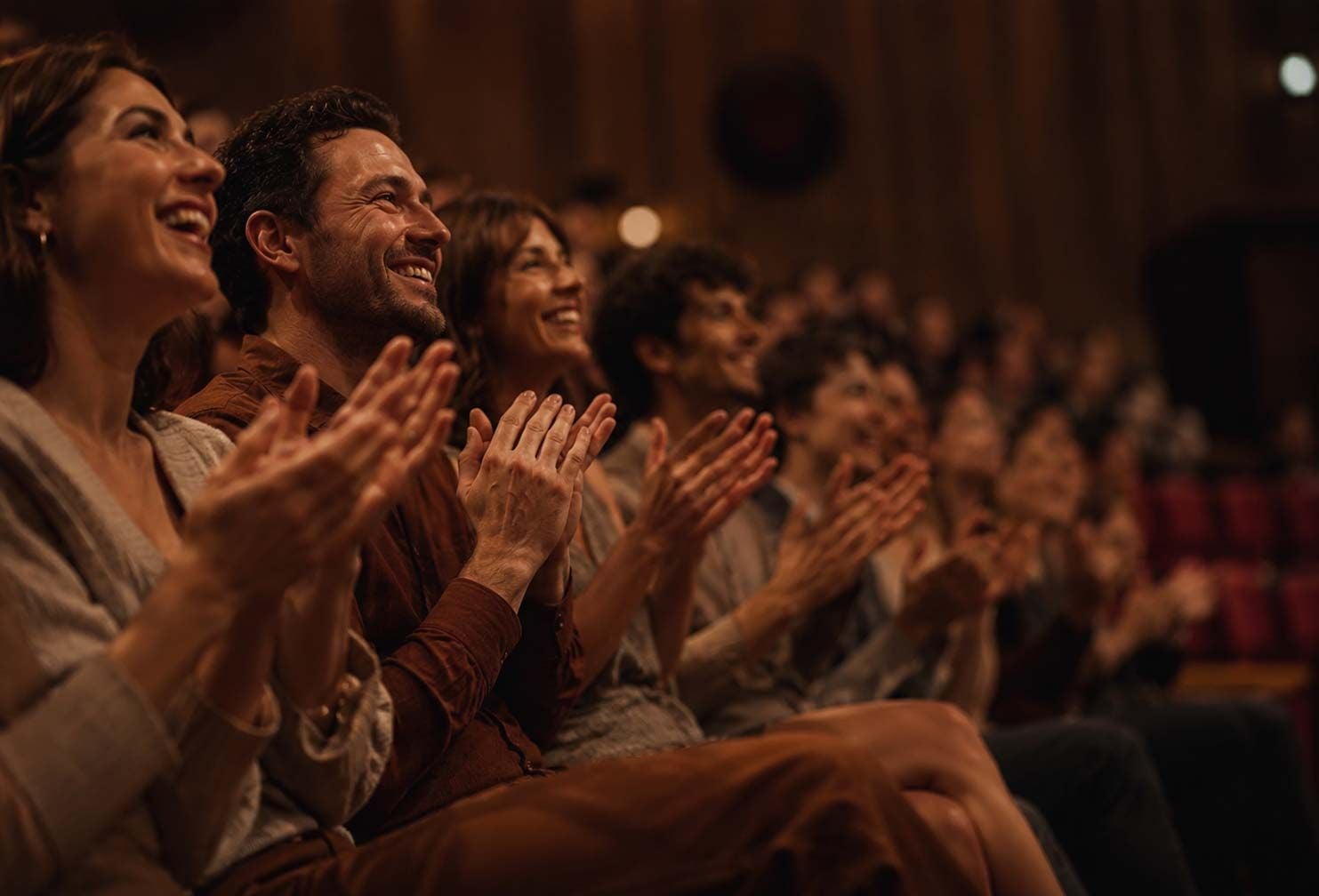 An audience of happy people, smiling and clapping enthusiastically in a dimly lit theater.