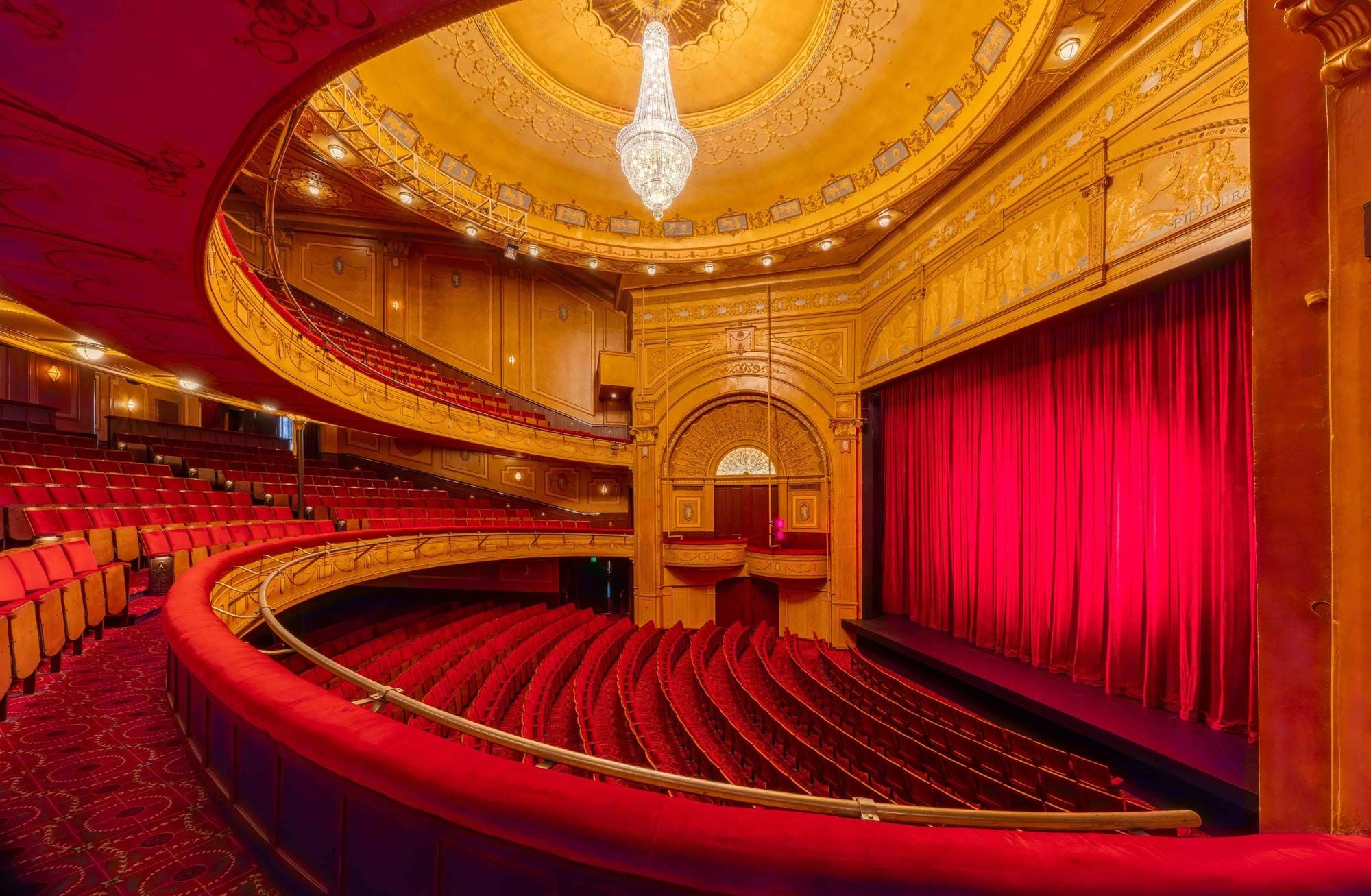 Opulent theater interior featuring red velvet seats, grand stage, and sparkling chandelier.