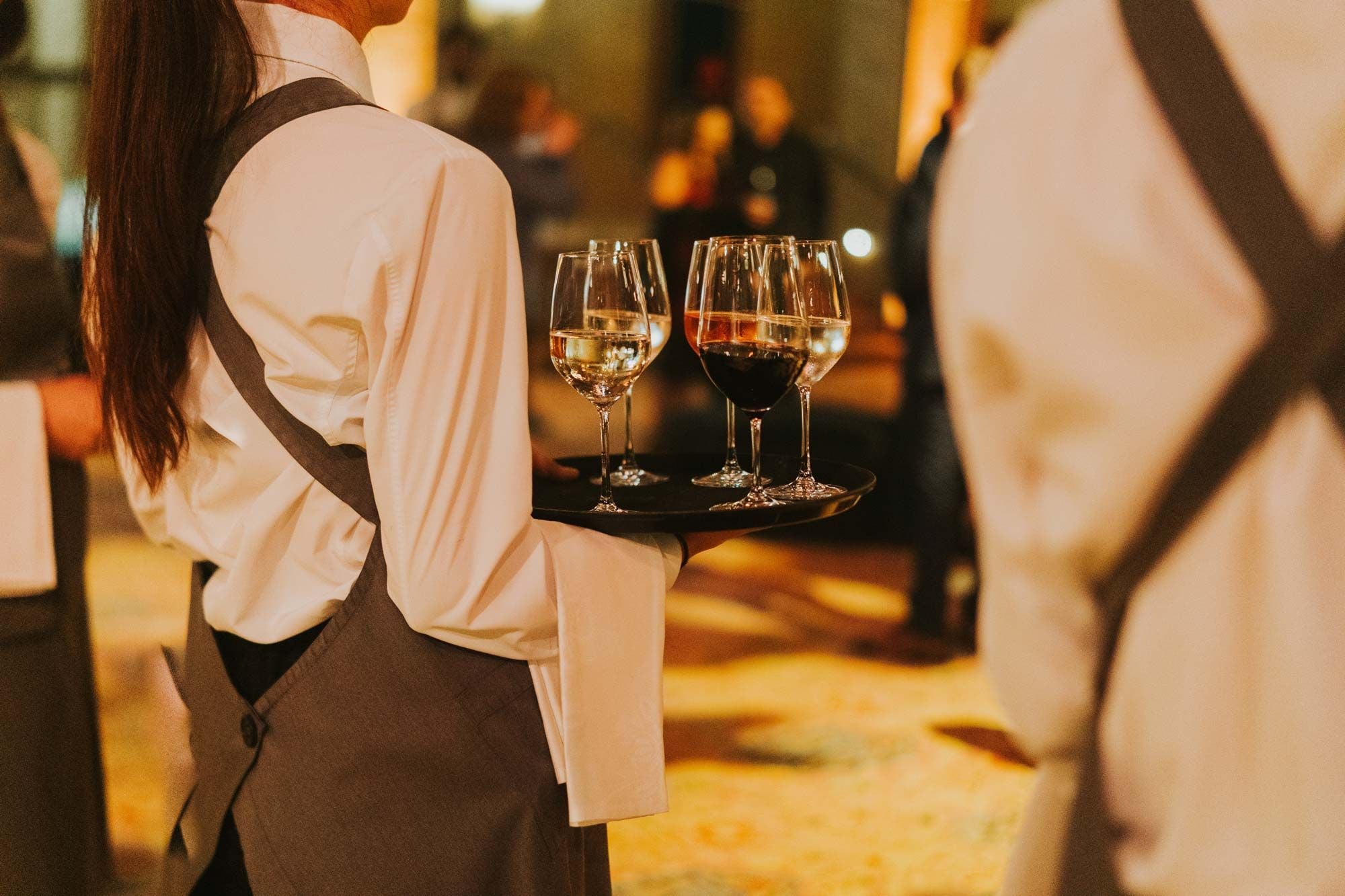Server wearing a white shirt and grey apron carrying a tray with wine glasses.
