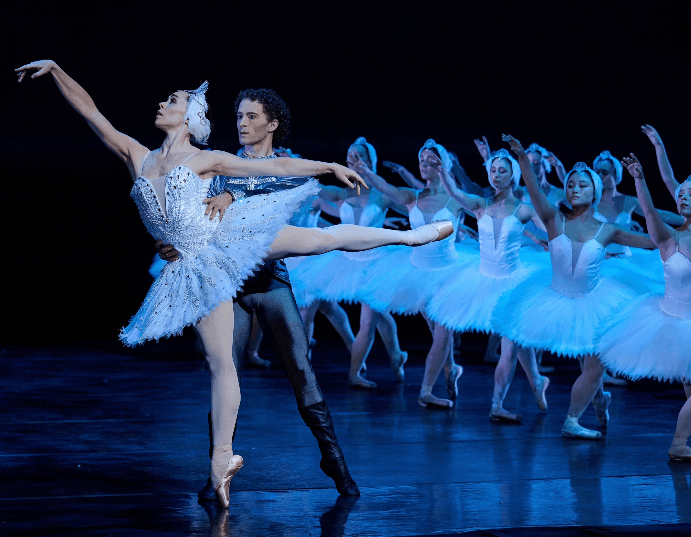 A ballerina in a white swan costume dances with a male partner and corps de ballet.