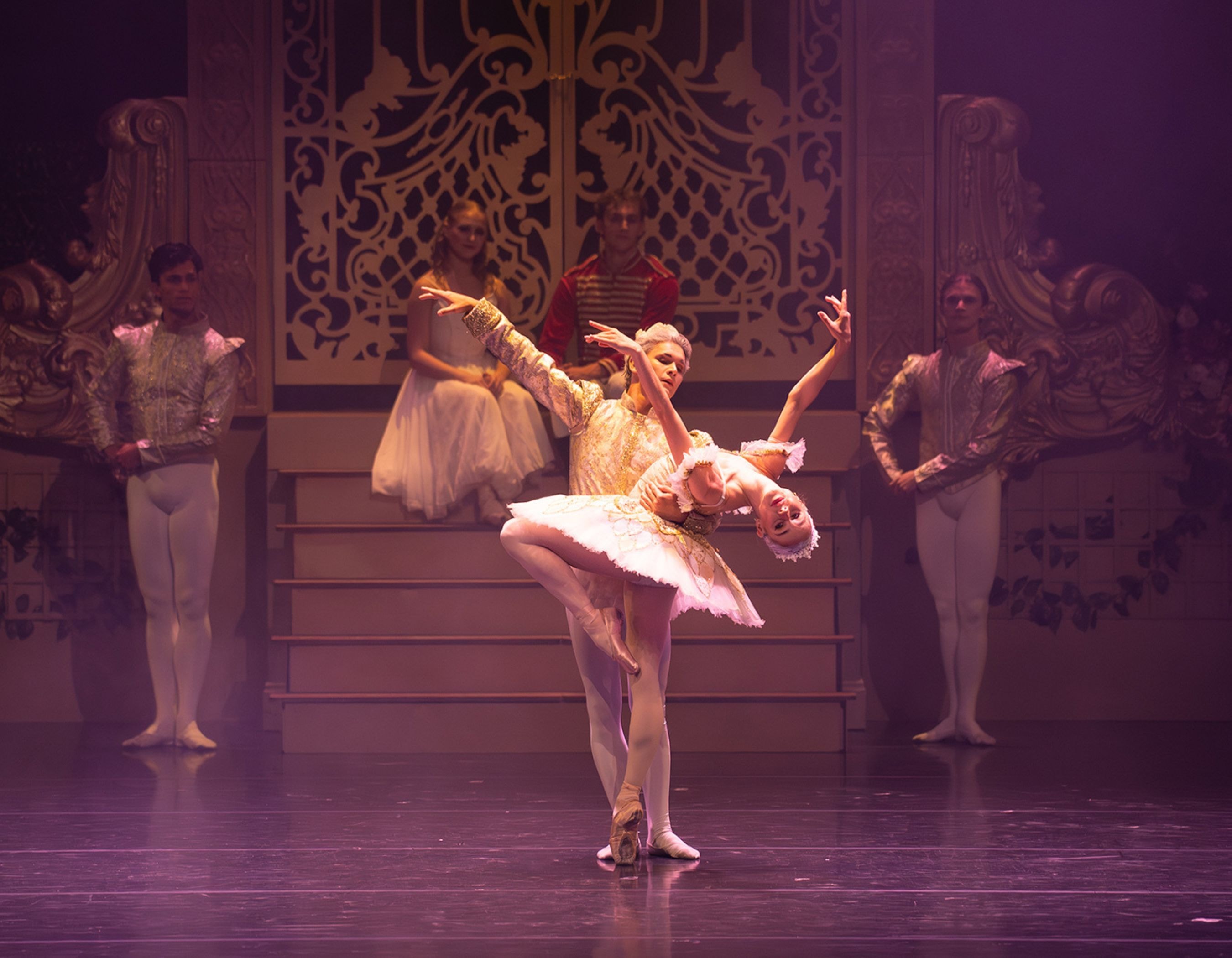 Two ballet dancers perform a graceful lift on stage with elaborate set pieces in the background.