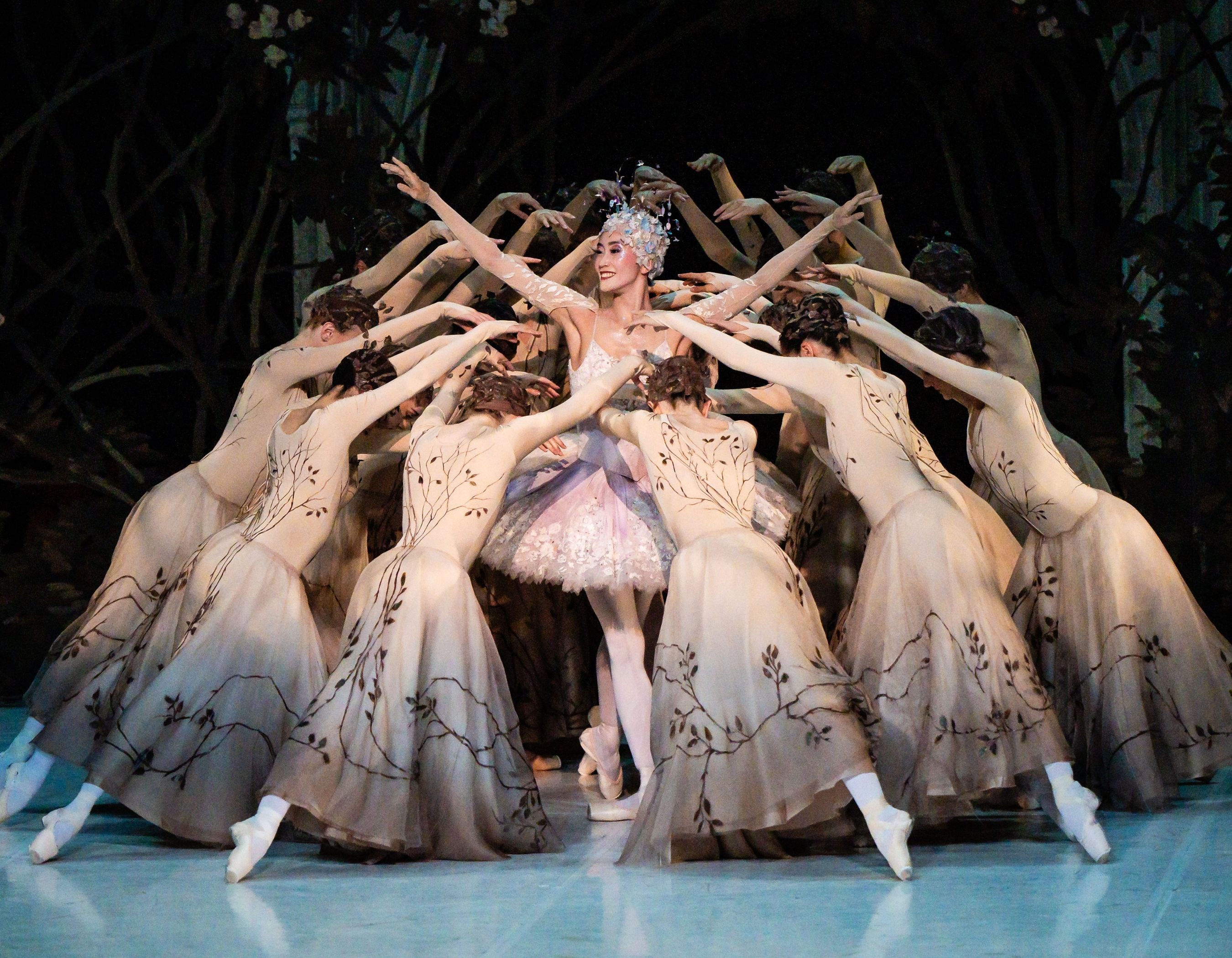 Prima ballerina in tiara surrounded by dancers with outstretched arms on a dark stage.