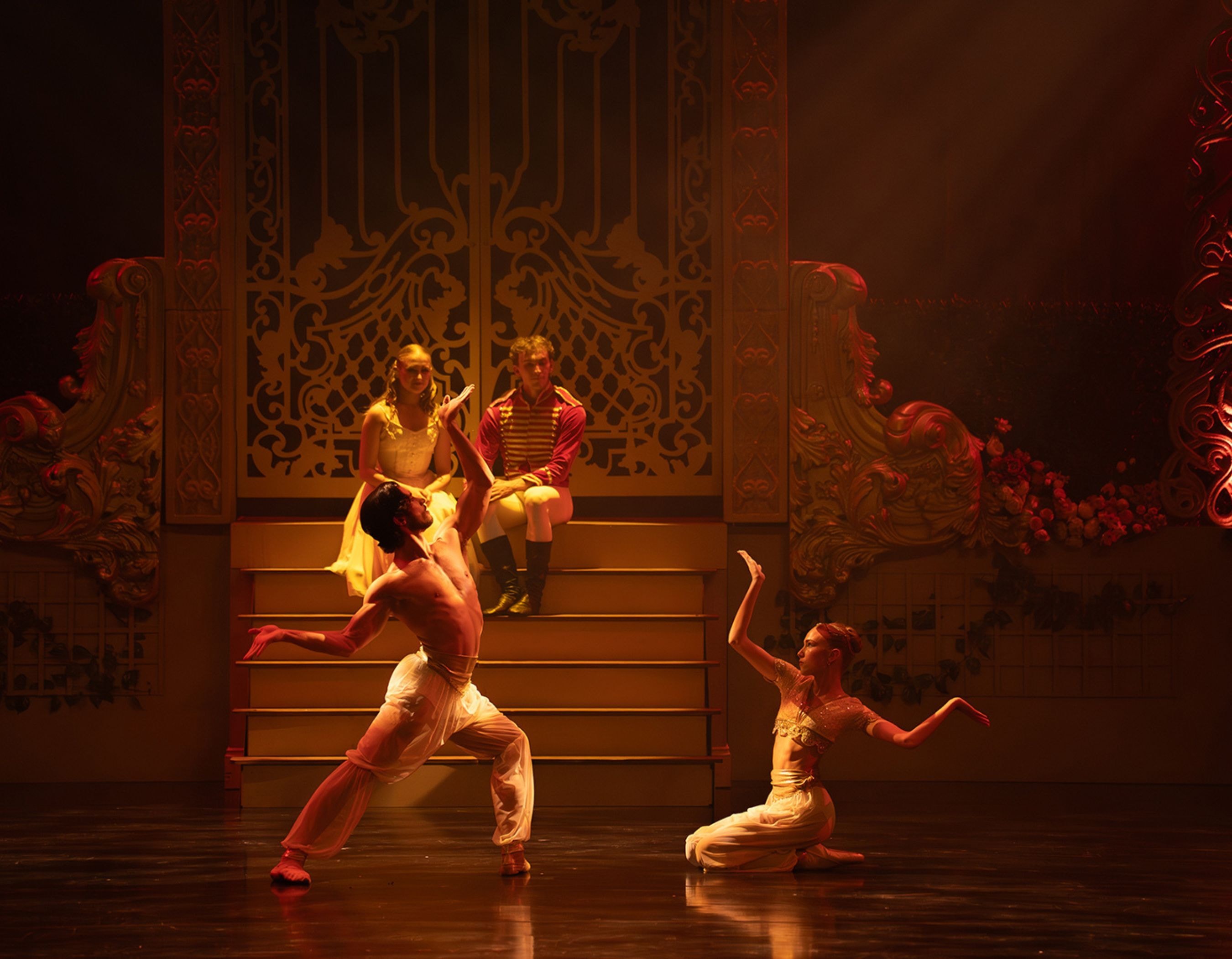 Two dancers perform on stage with two seated figures in the background.