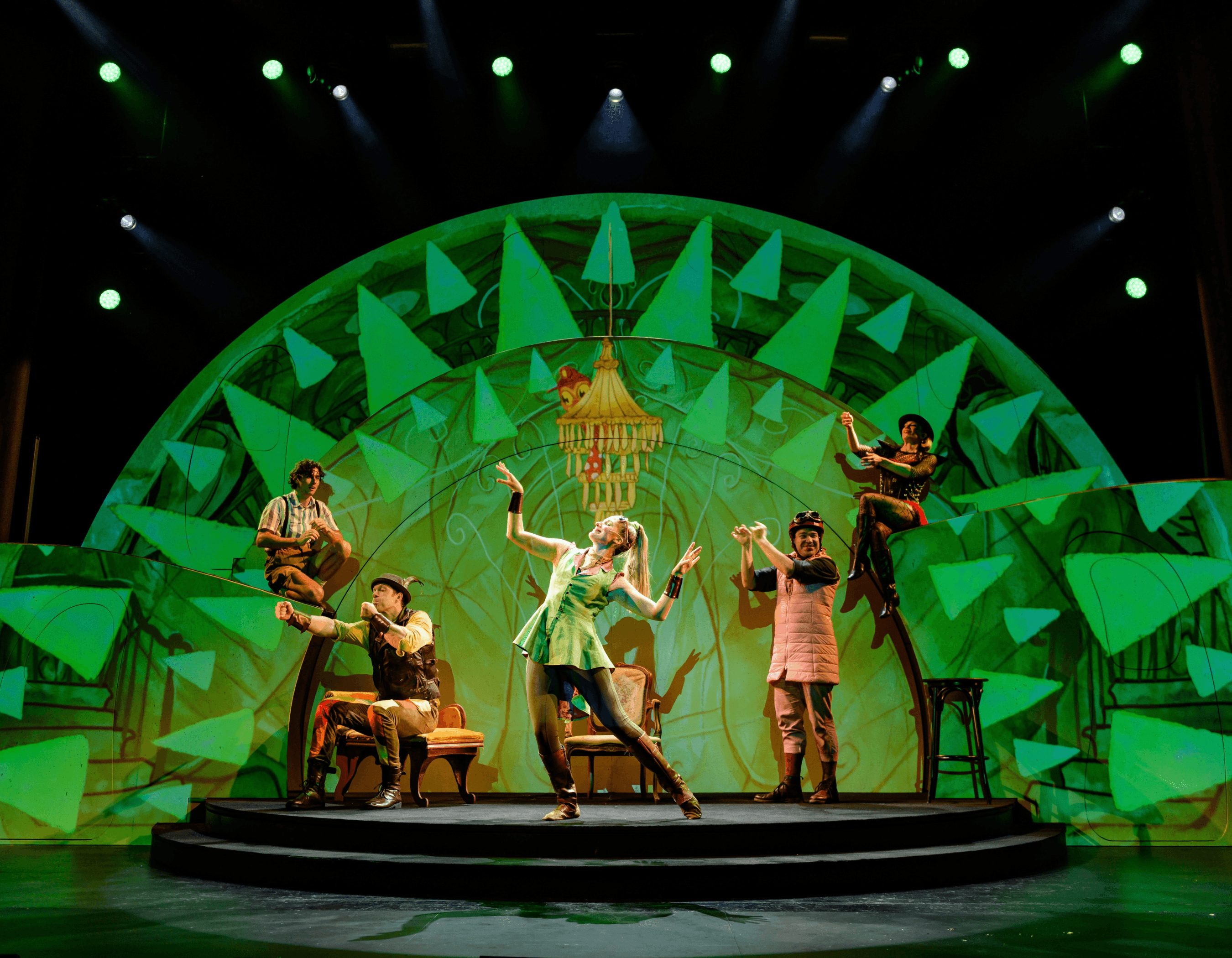 Performers in costumes dance on a brightly lit stage with a vibrant green geometric backdrop.