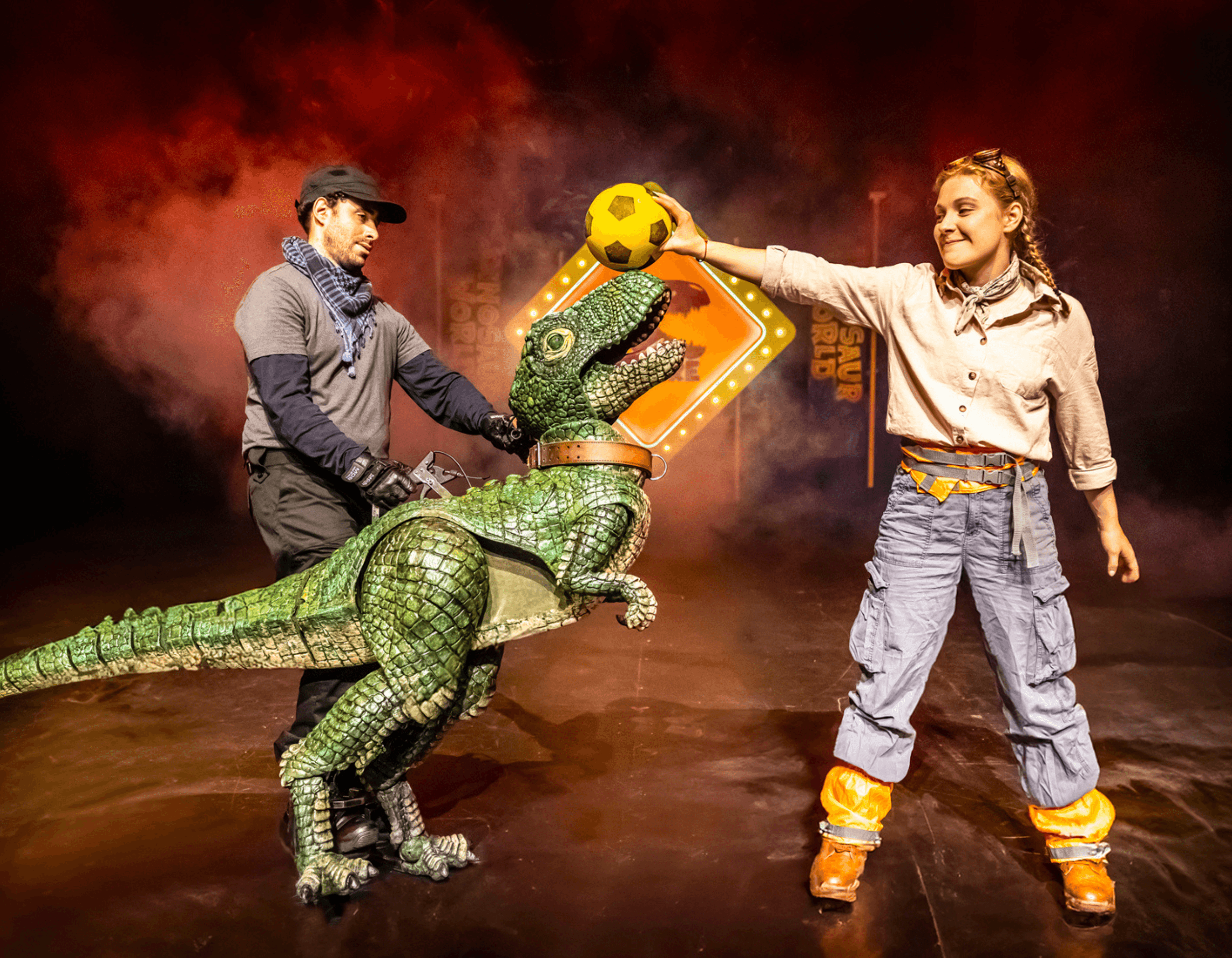 Two performers interact with a realistic green dinosaur puppet on a dimly lit stage.