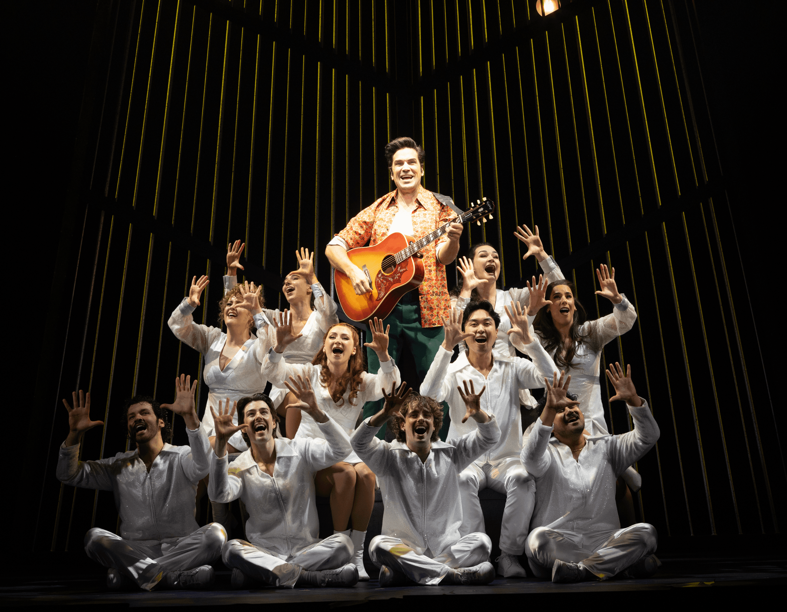 A theatrical ensemble performs on stage, a central figure playing guitar surrounded by enthusiastic singers.
