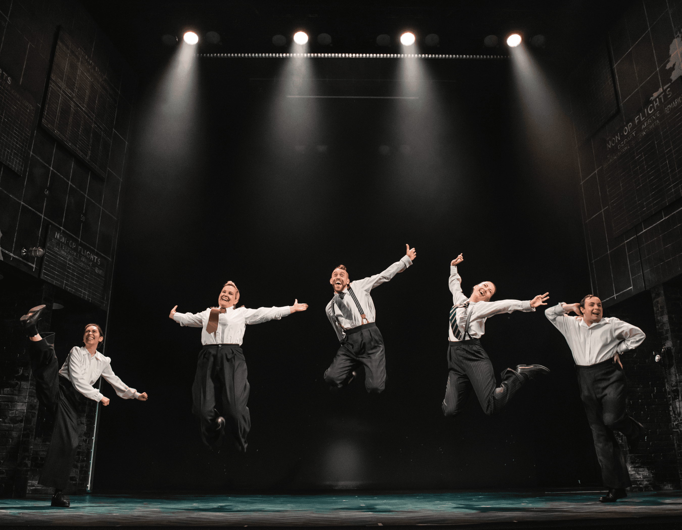 Energetic performers leap in unison on a theatrical stage under bright spotlights.