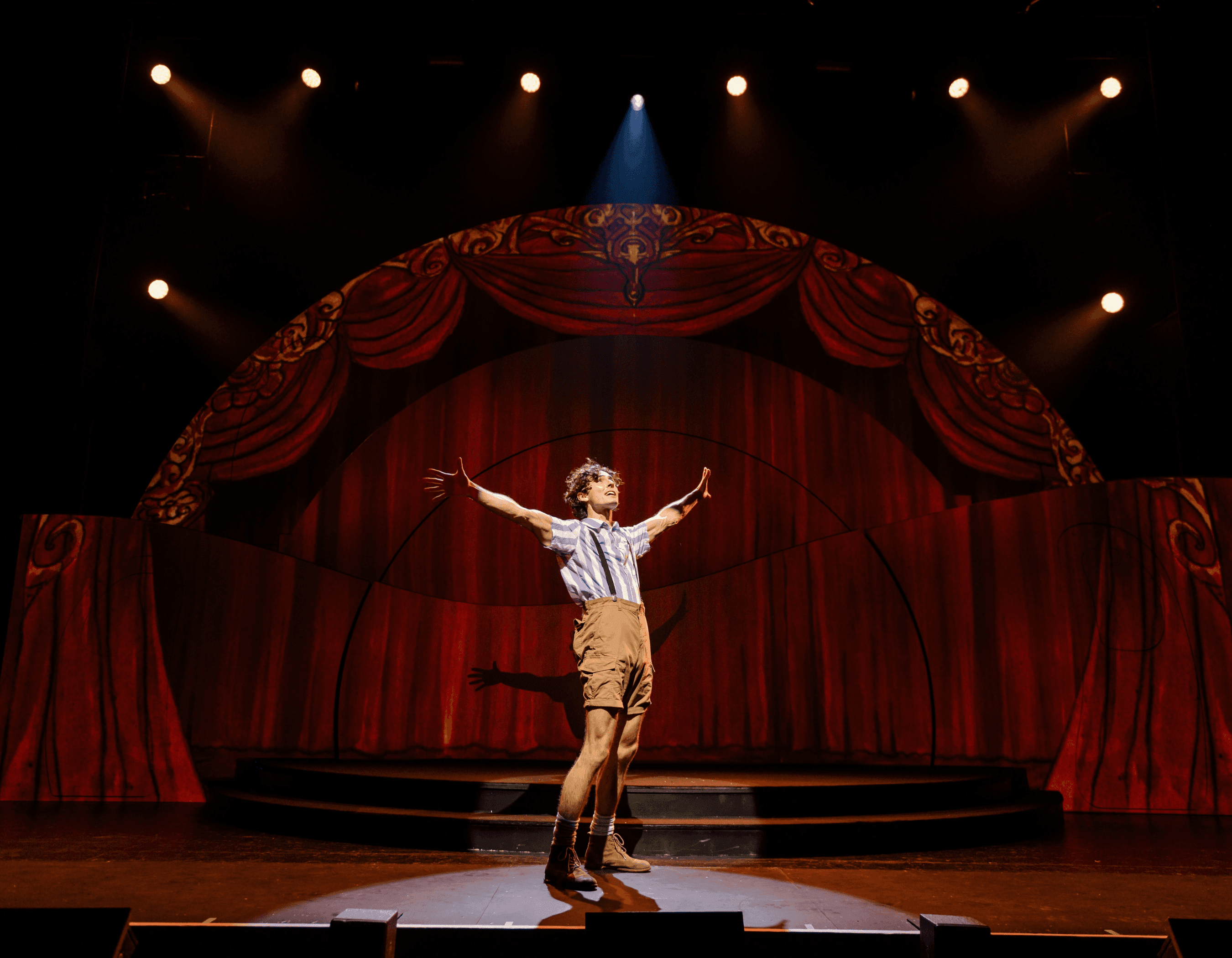 Performer on a theater stage in a spotlight with arms outstretched before red curtains.