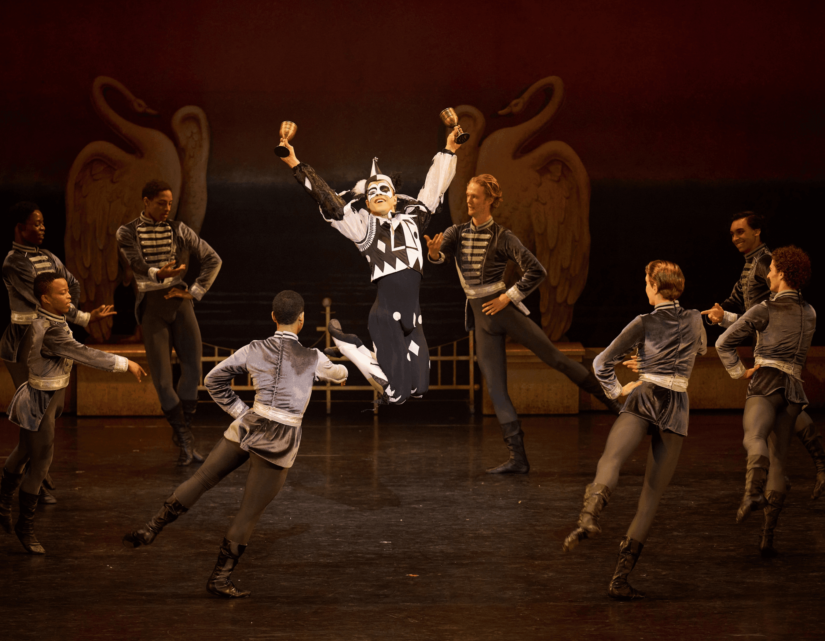 Harlequin dancer leaps joyfully with chalices, surrounded by ballet dancers on stage.