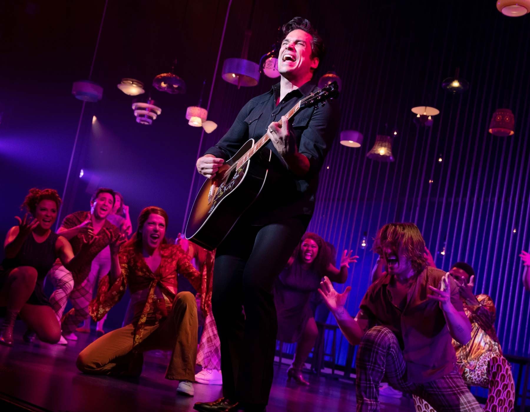 Singer in black shirt playing guitar and singing on stage with dancers.