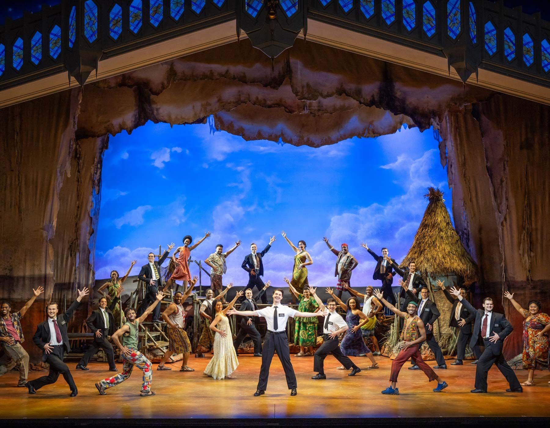 A large ensemble of performers in diverse costumes dances energetically on a vibrant stage.