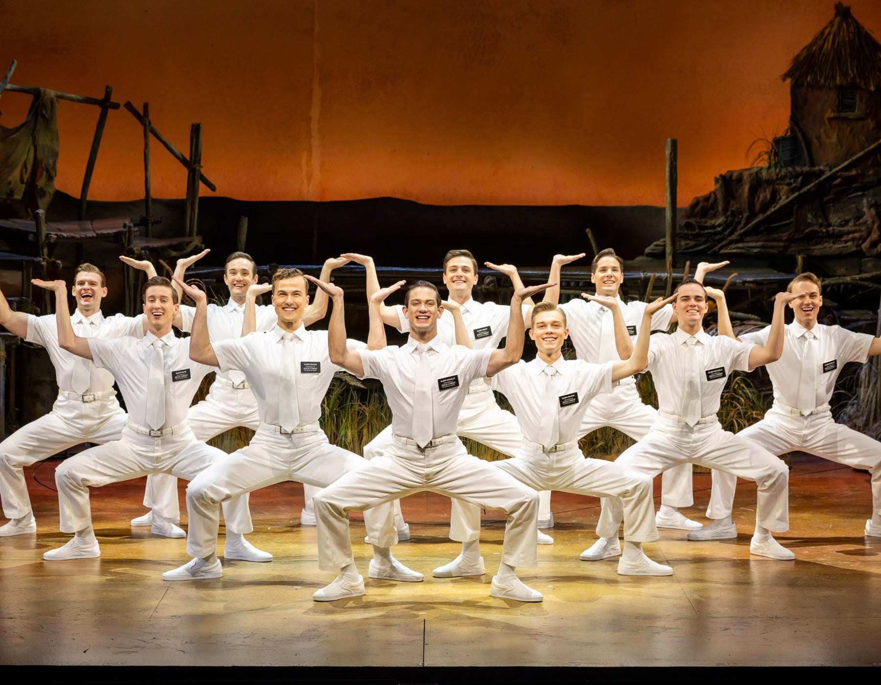 Eleven men in white shirts and pants perform a synchronized, angular dance pose.