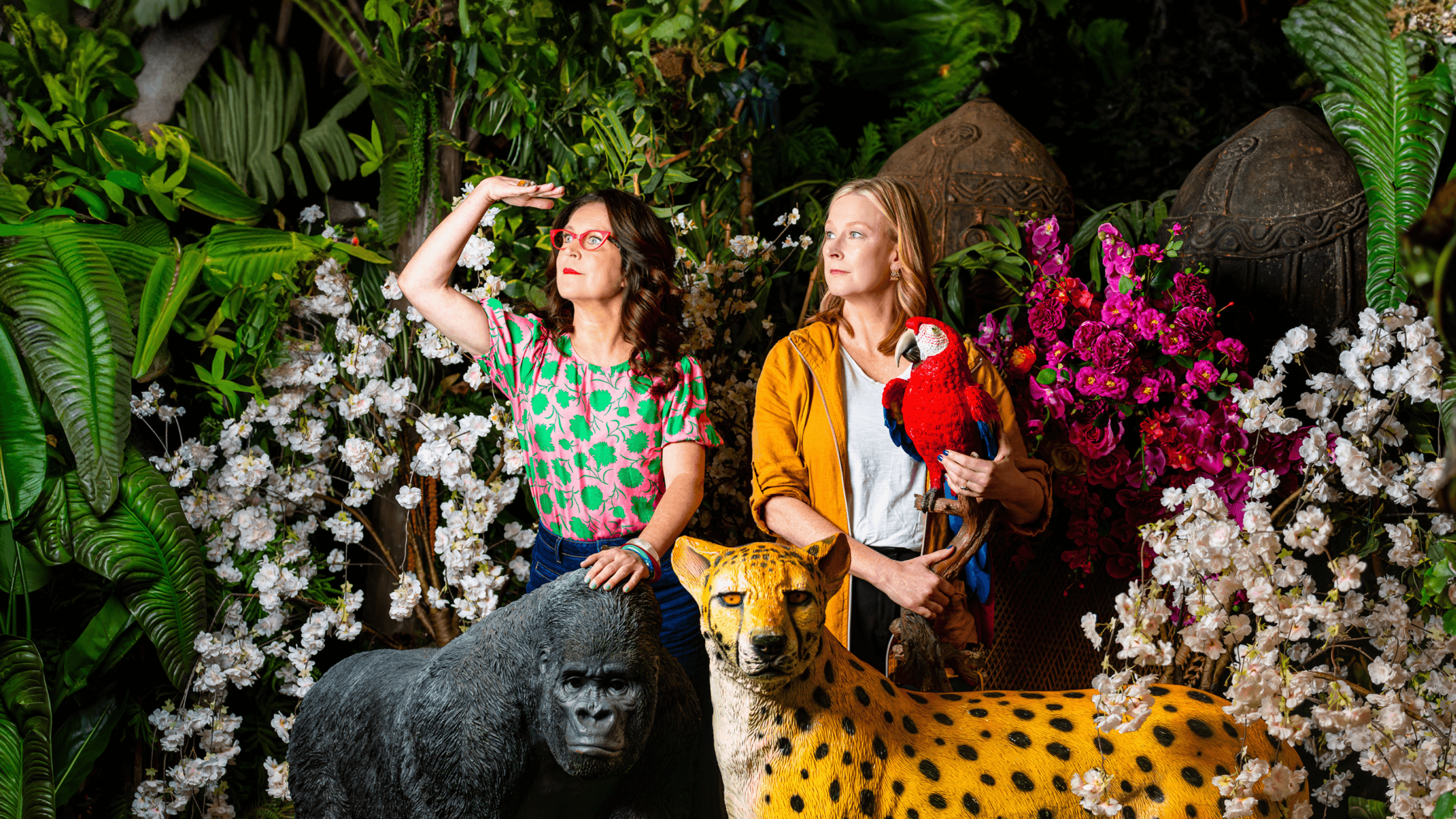 Two women, one with glasses, in a lush jungle with a parrot and animal statues.