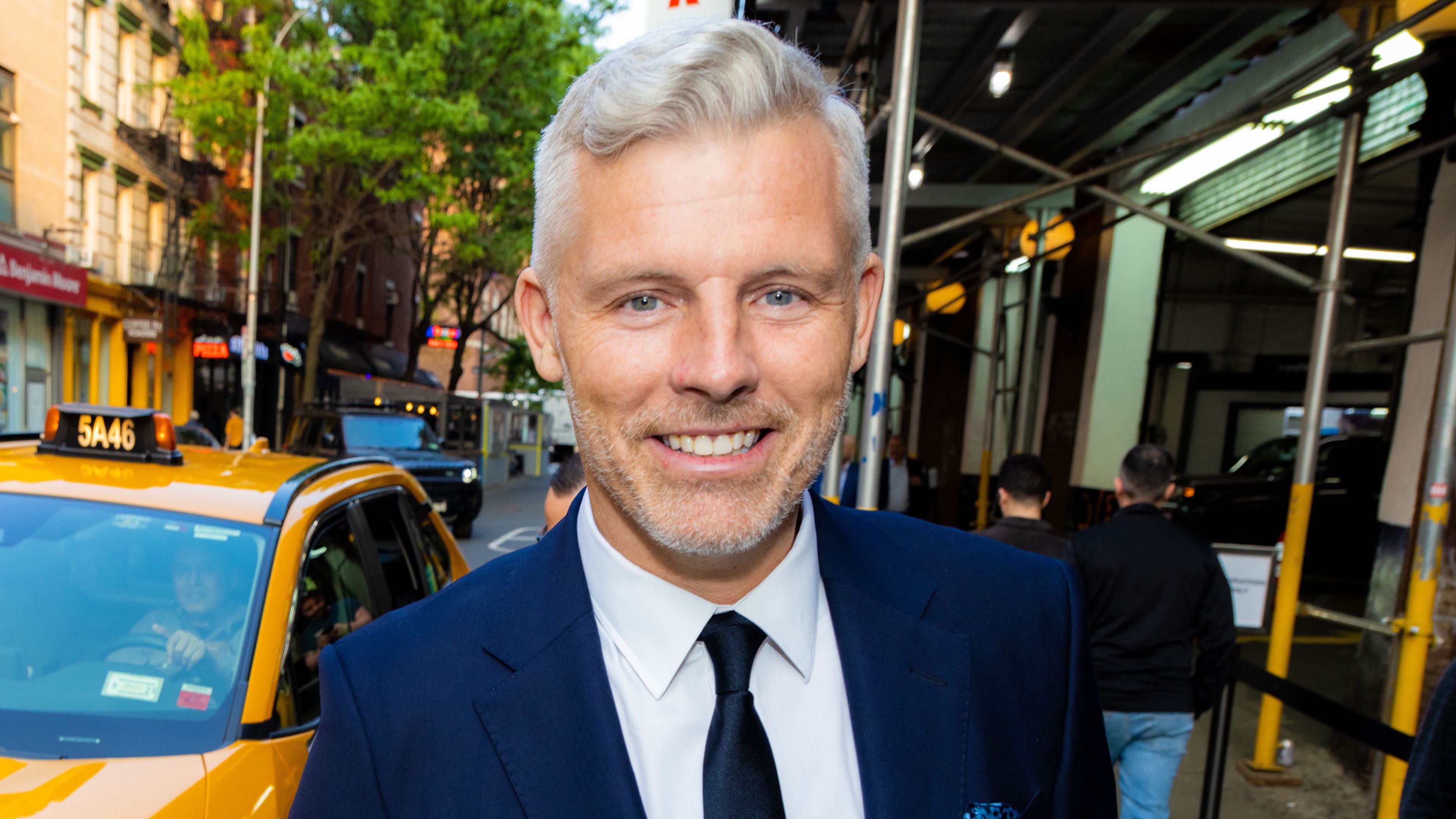 Smiling man with grey hair and beard in a navy suit on a city street.