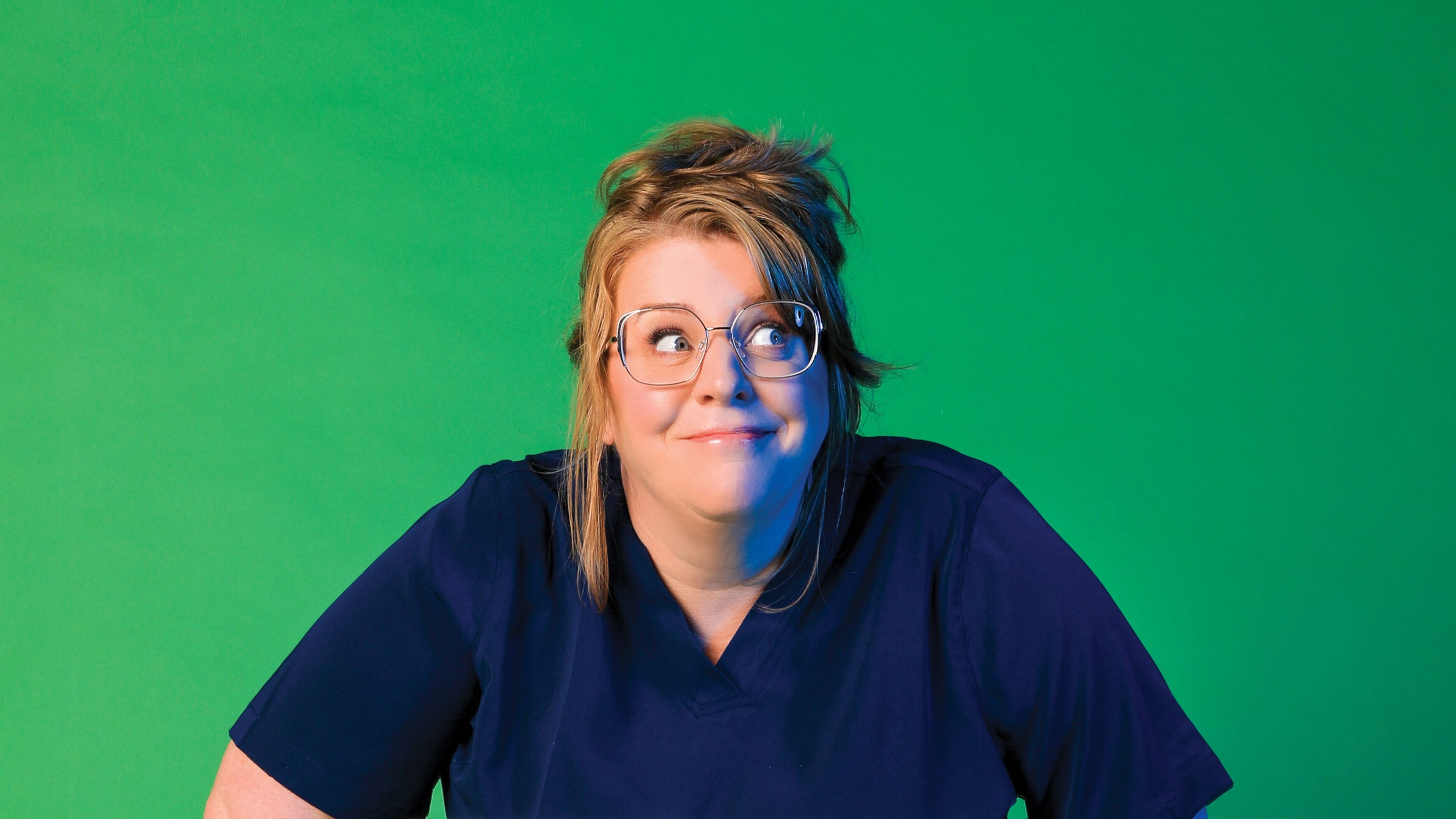 Woman with glasses and humorous expression wearing navy scrubs against a green background.