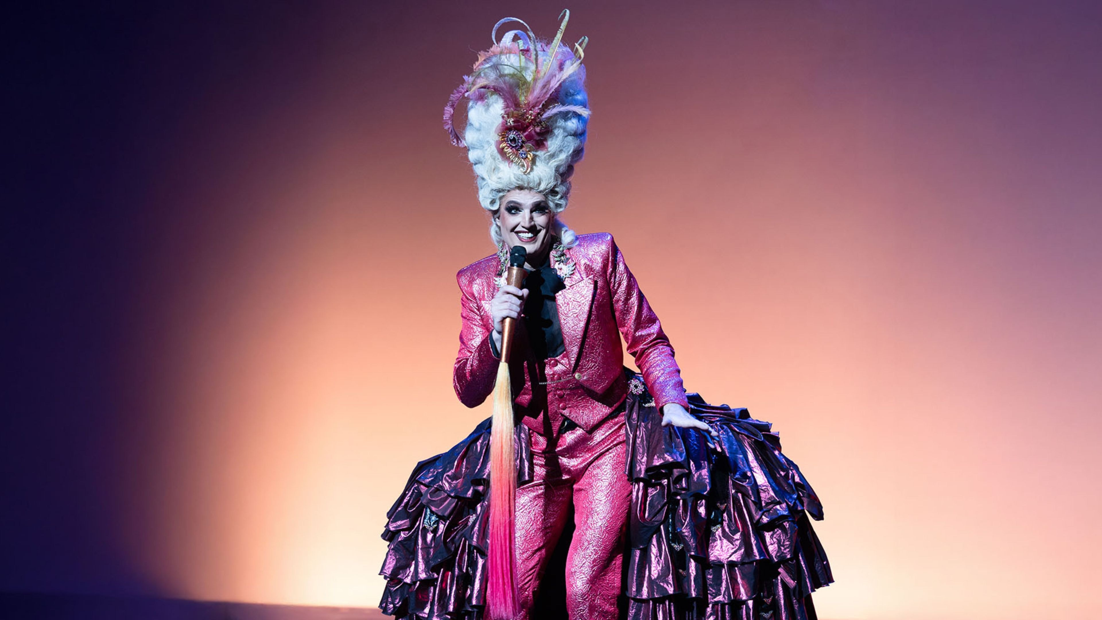 Drag performer in shiny pink suit, elaborate wig, and ruffled skirt holds a microphone.