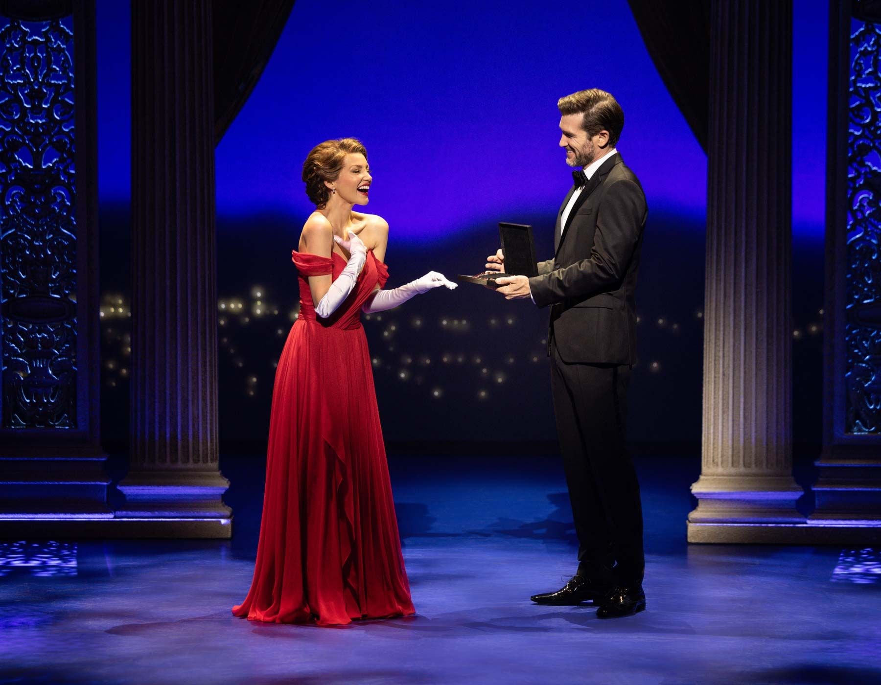 Man in tuxedo offers hat to a woman in a red gown on stage.