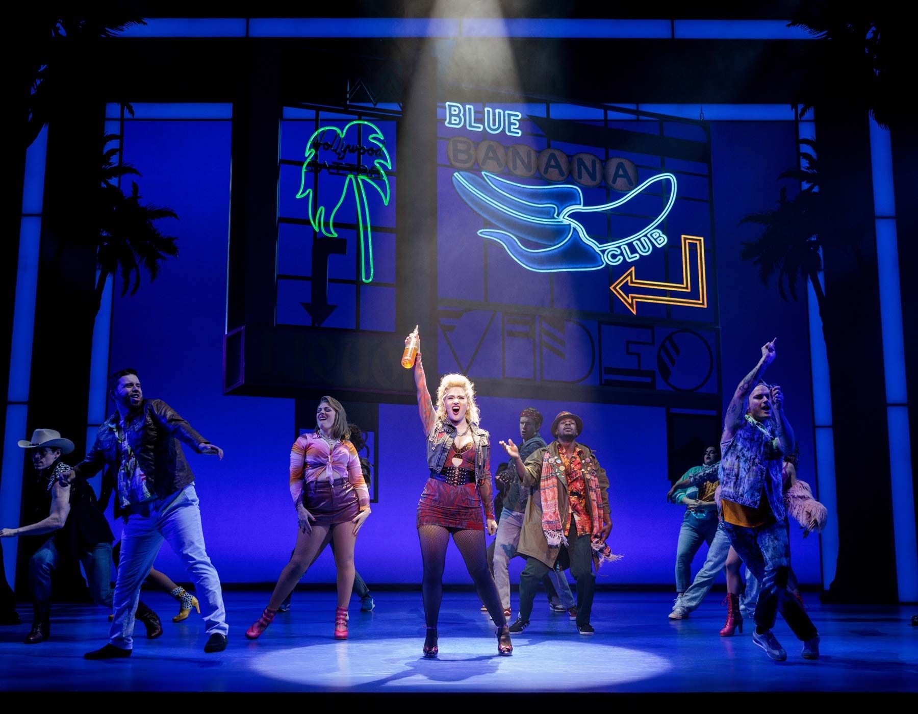 Woman in red sequin outfit sings, surrounded by dancers under blue neon signs.