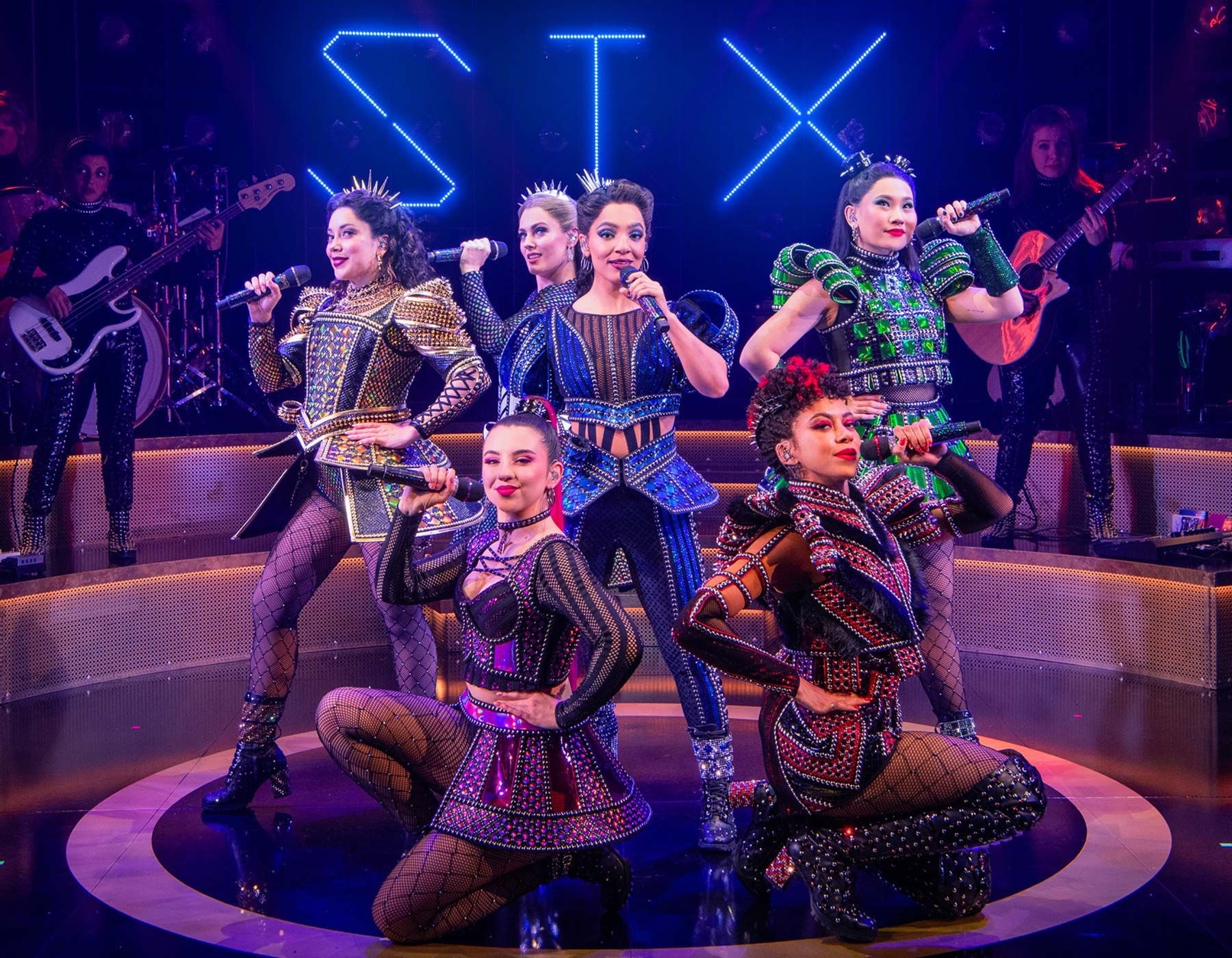 Six women in ornate, modern historical costumes pose dynamically on a stage.