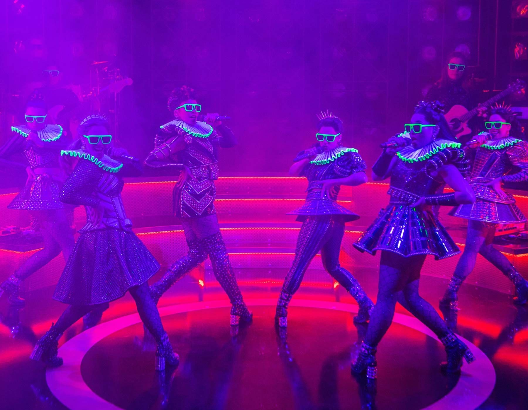 Six performers in futuristic outfits with glowing accessories dance under purple blacklight.