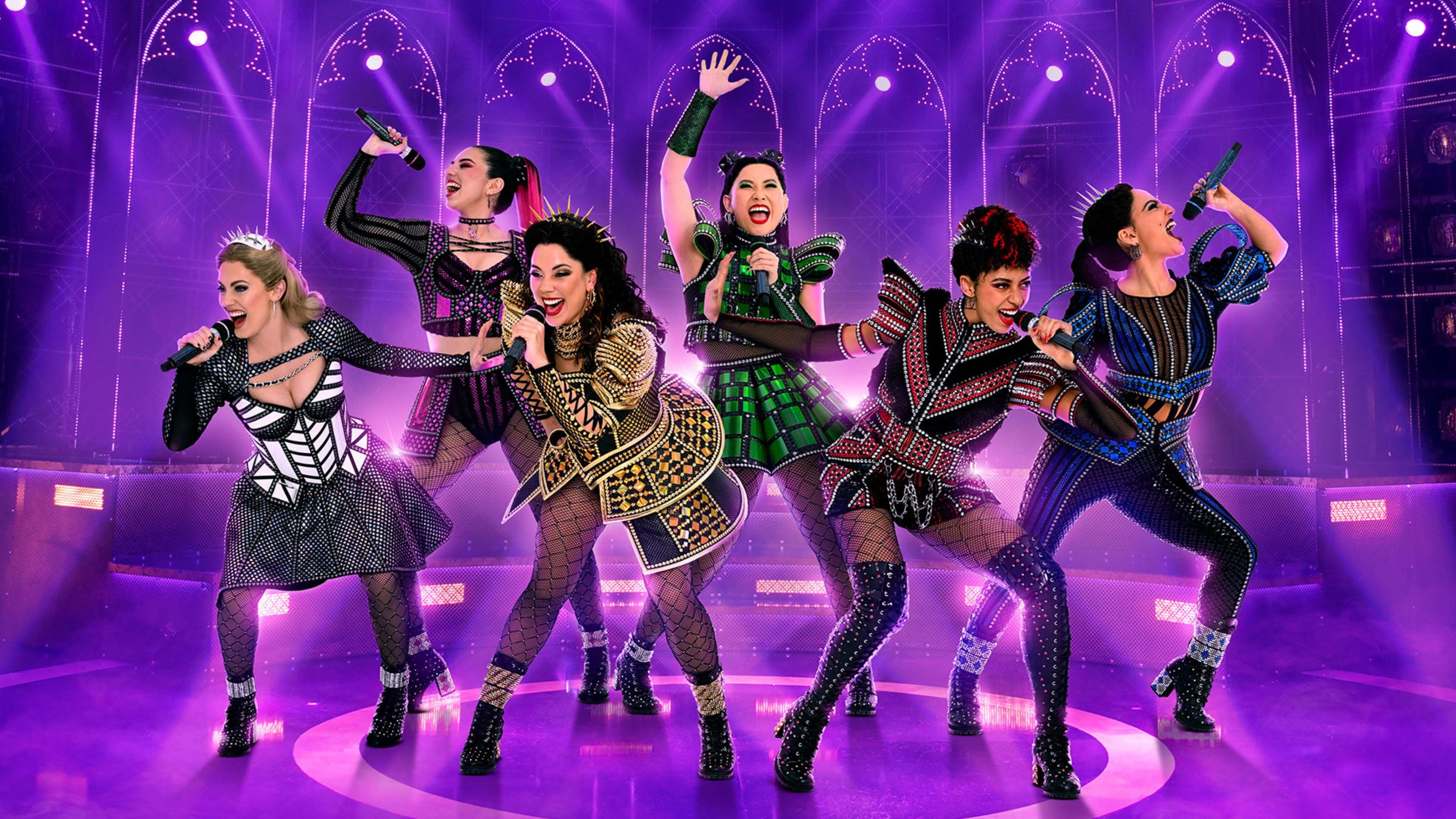 Six female performers in vibrant, modern queen costumes sing on a purple stage.