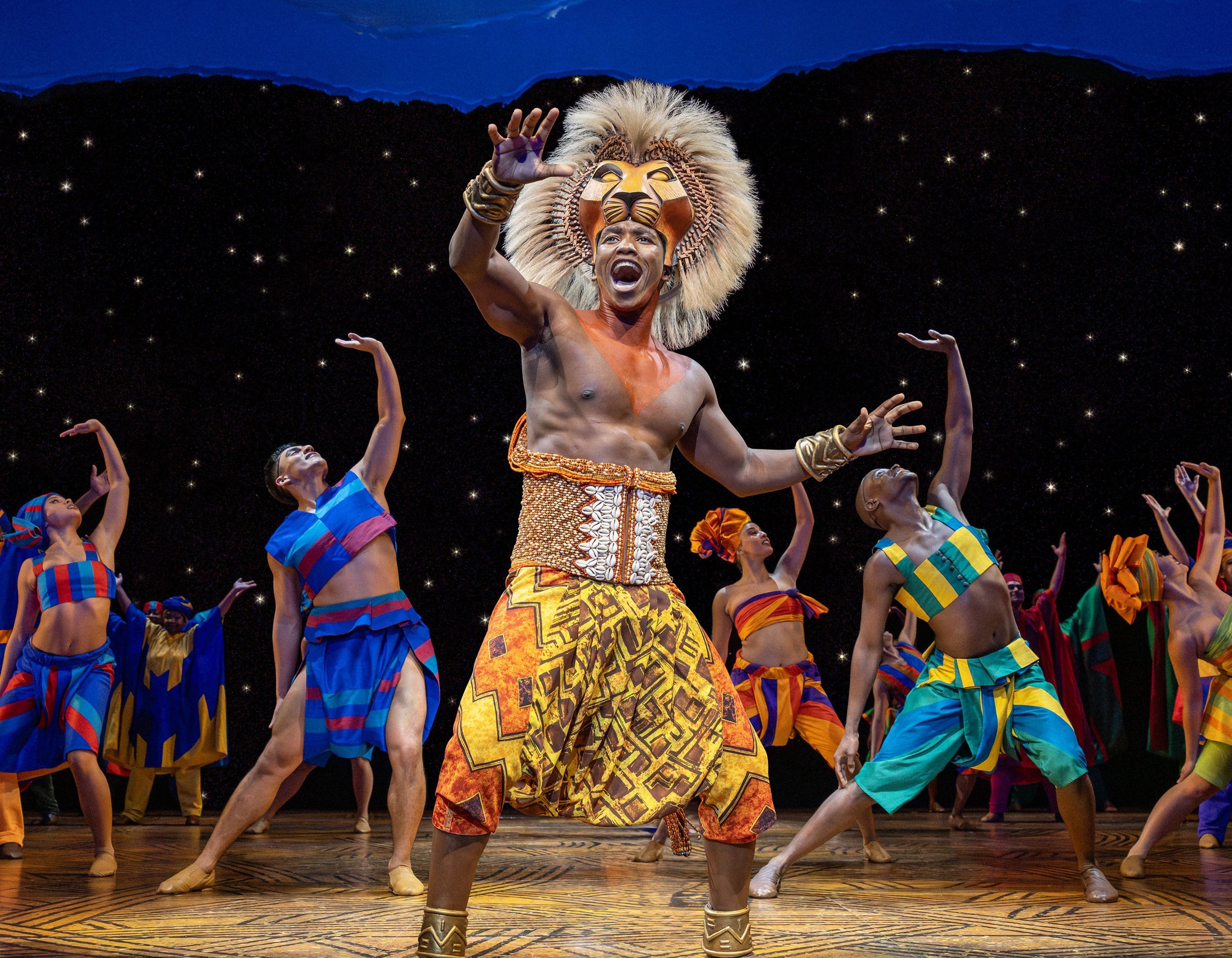 Performer as Simba with lion mask and costume on stage surrounded by dancers.