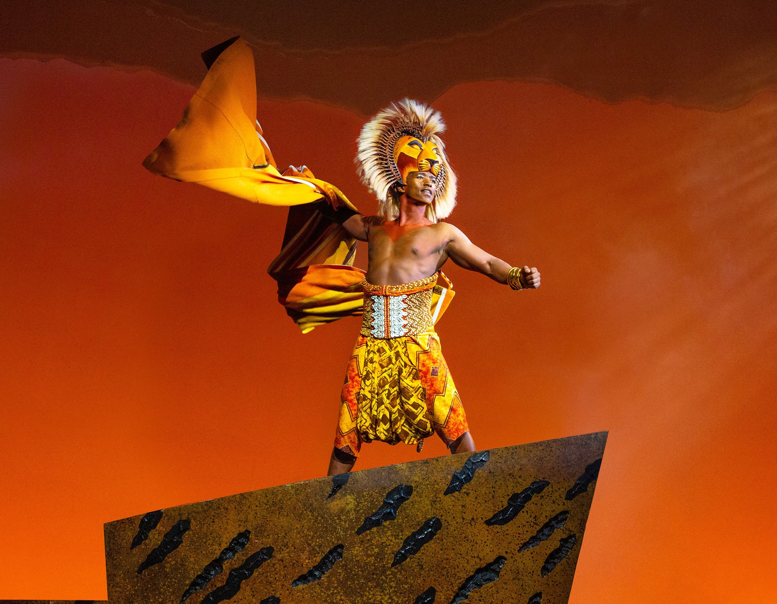 Performer in an elaborate lion costume stands on a stage rock against an orange background.
