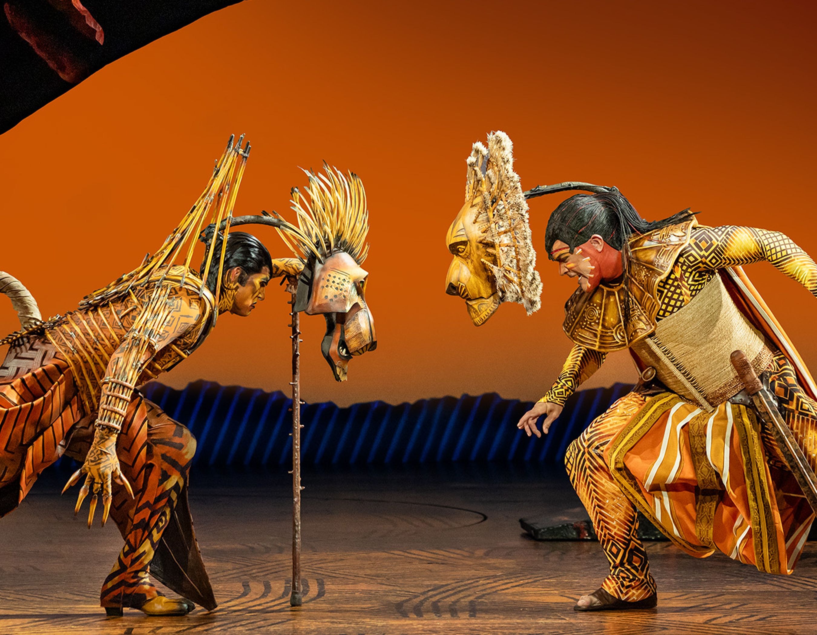 Two performers in elaborate animal costumes and masks face each other on stage.