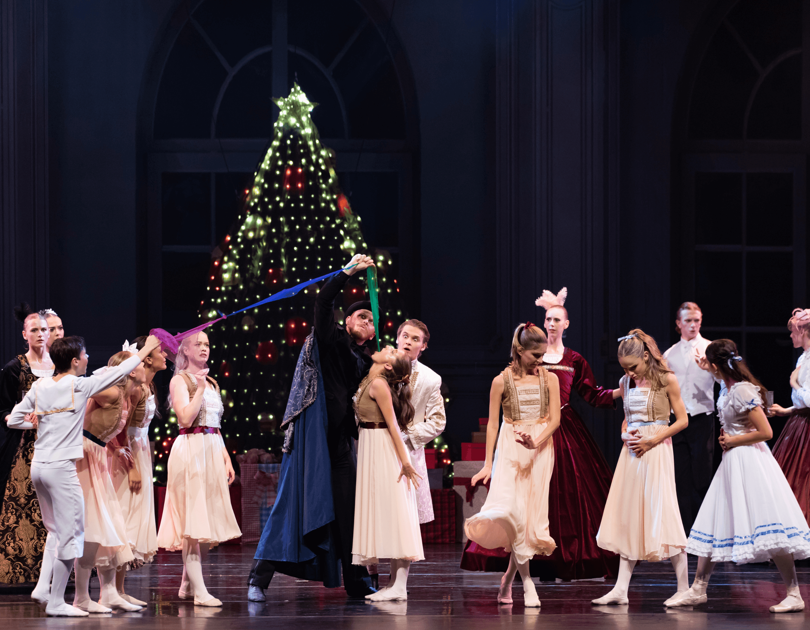 Ballet dancers perform on stage with a large decorated Christmas tree in the background.