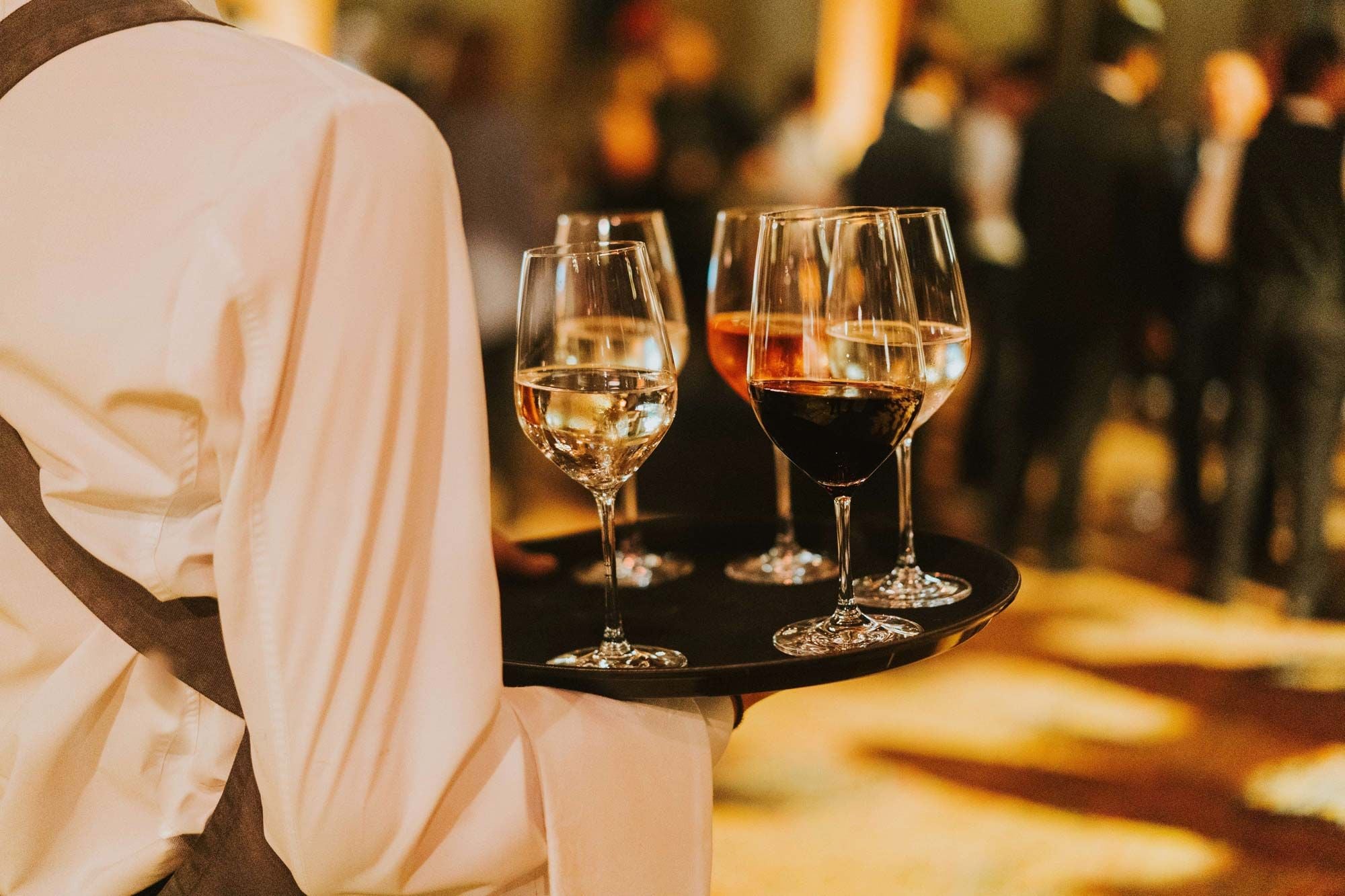 A waiter carries a black tray with glasses of red and white wine at an event.