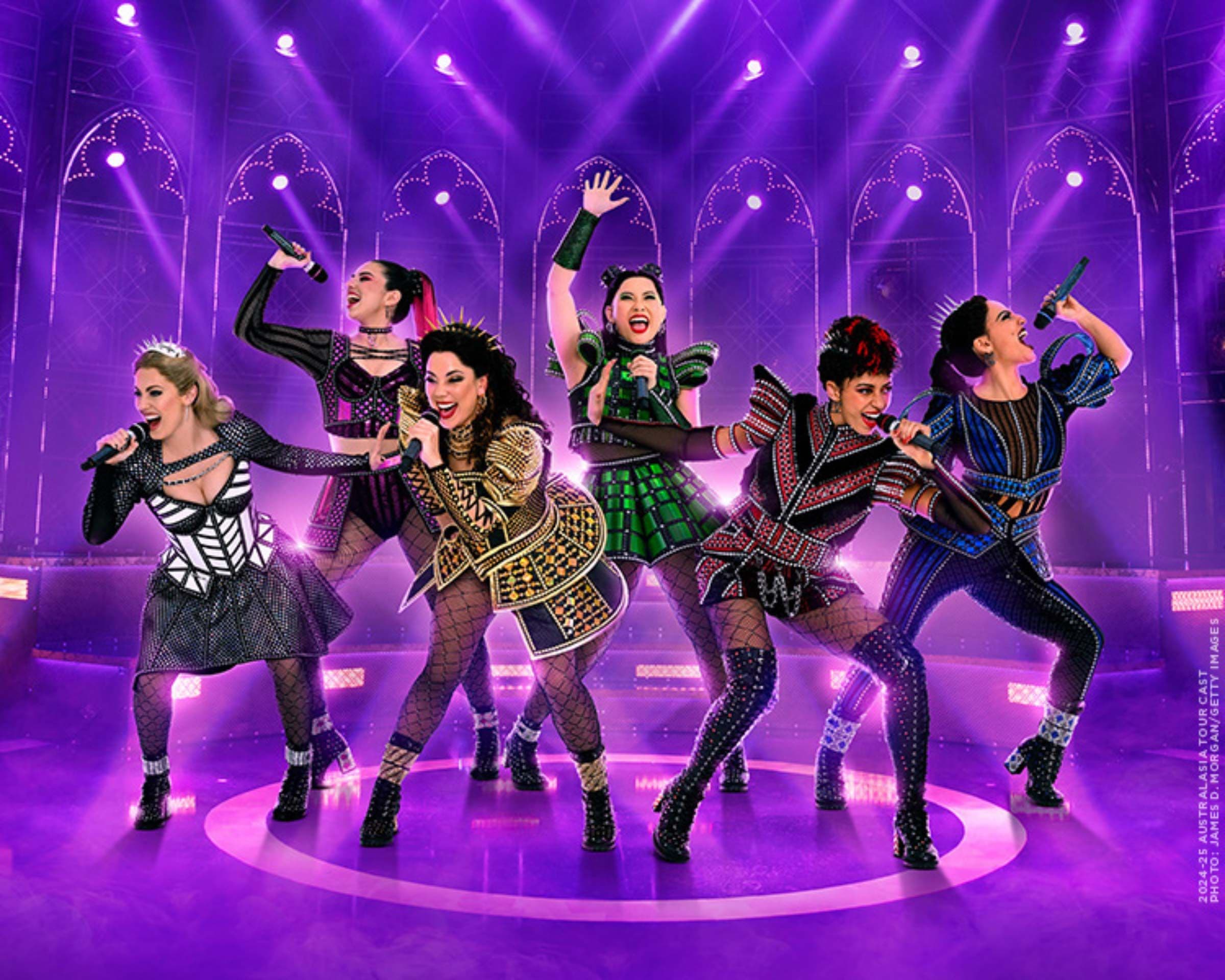 Six female performers in vibrant queen-inspired costumes sing on a purple stage with spotlights.