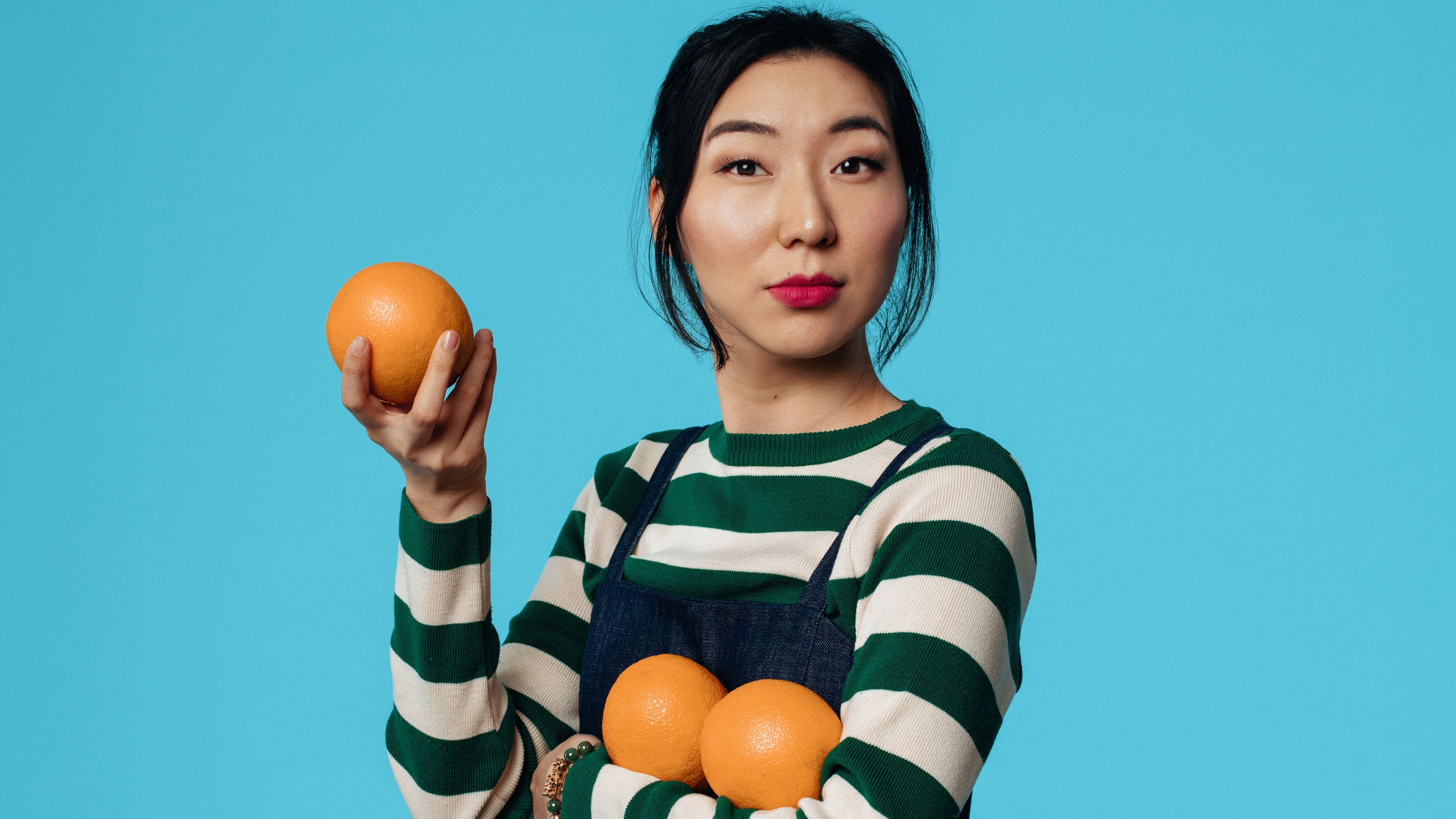 Young Asian woman with red lipstick holds oranges against a vibrant blue background.