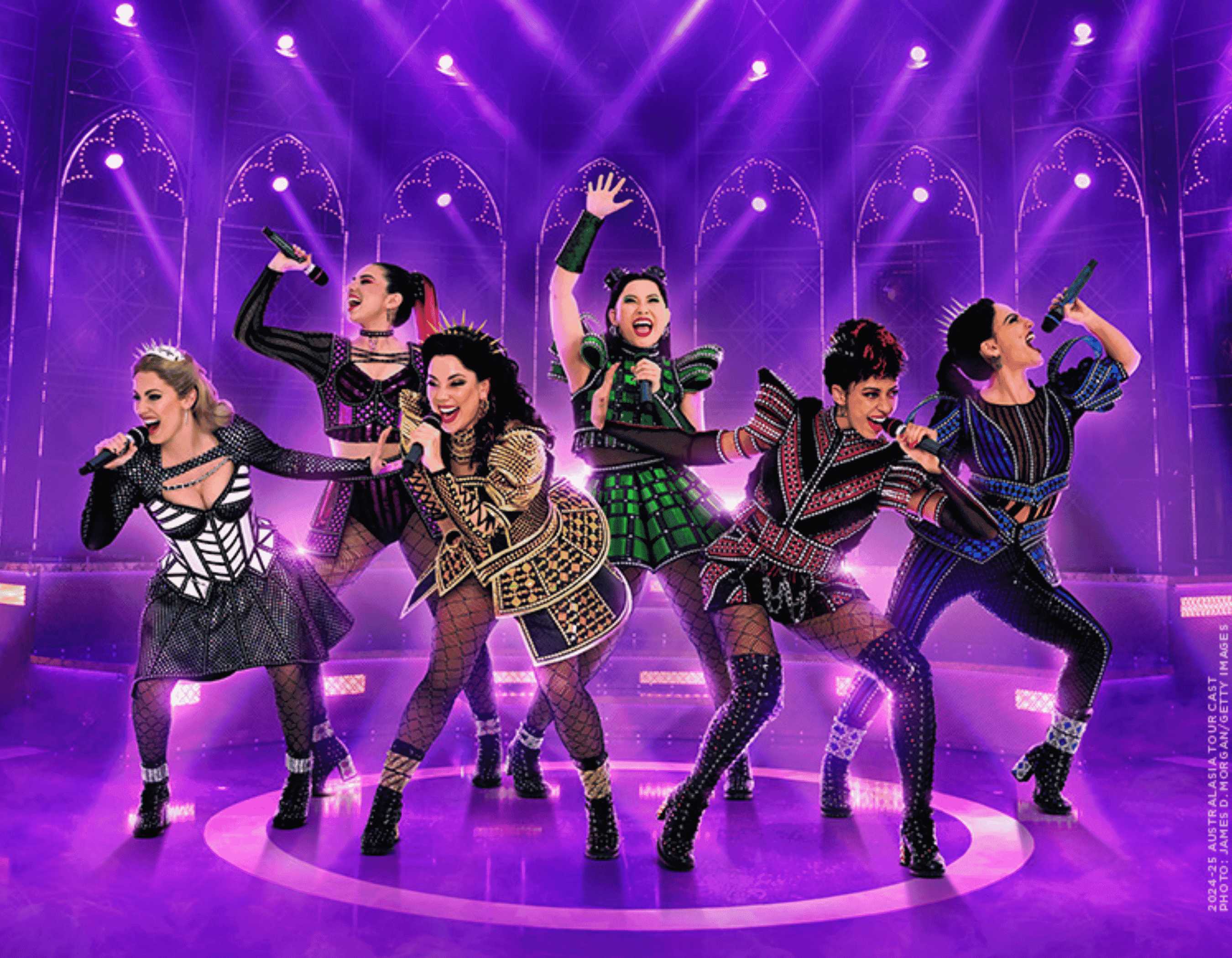 Six women in vibrant pop star costumes sing on a stage with microphones under purple lights.