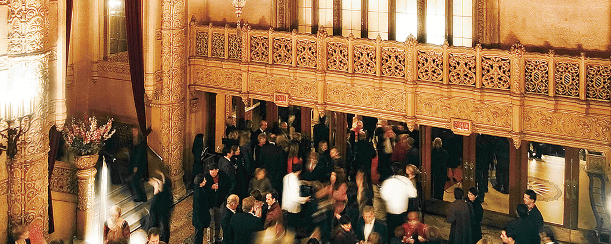 Crowd of people socializing in an opulent, multi-level theatre lobby with ornate architecture.
