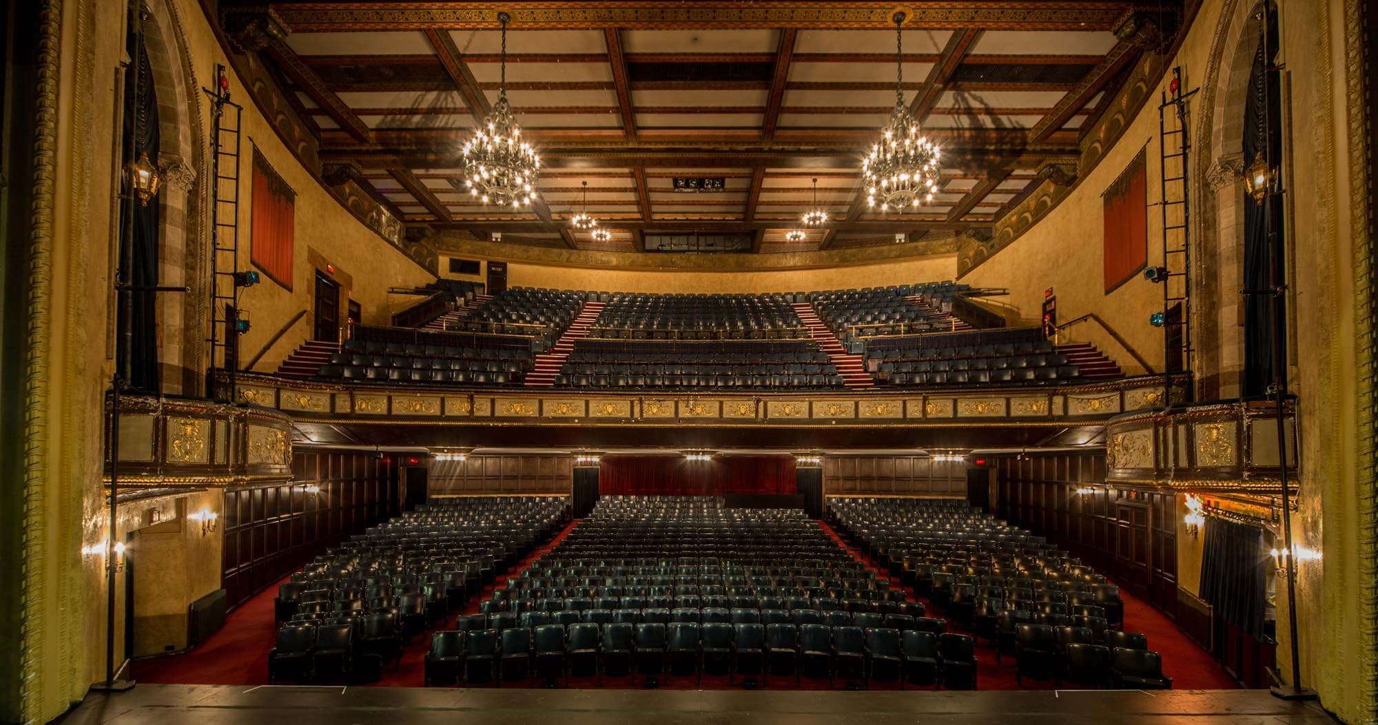 Ornate, empty theater with rows of dark seats, a balcony, and chandeliers.